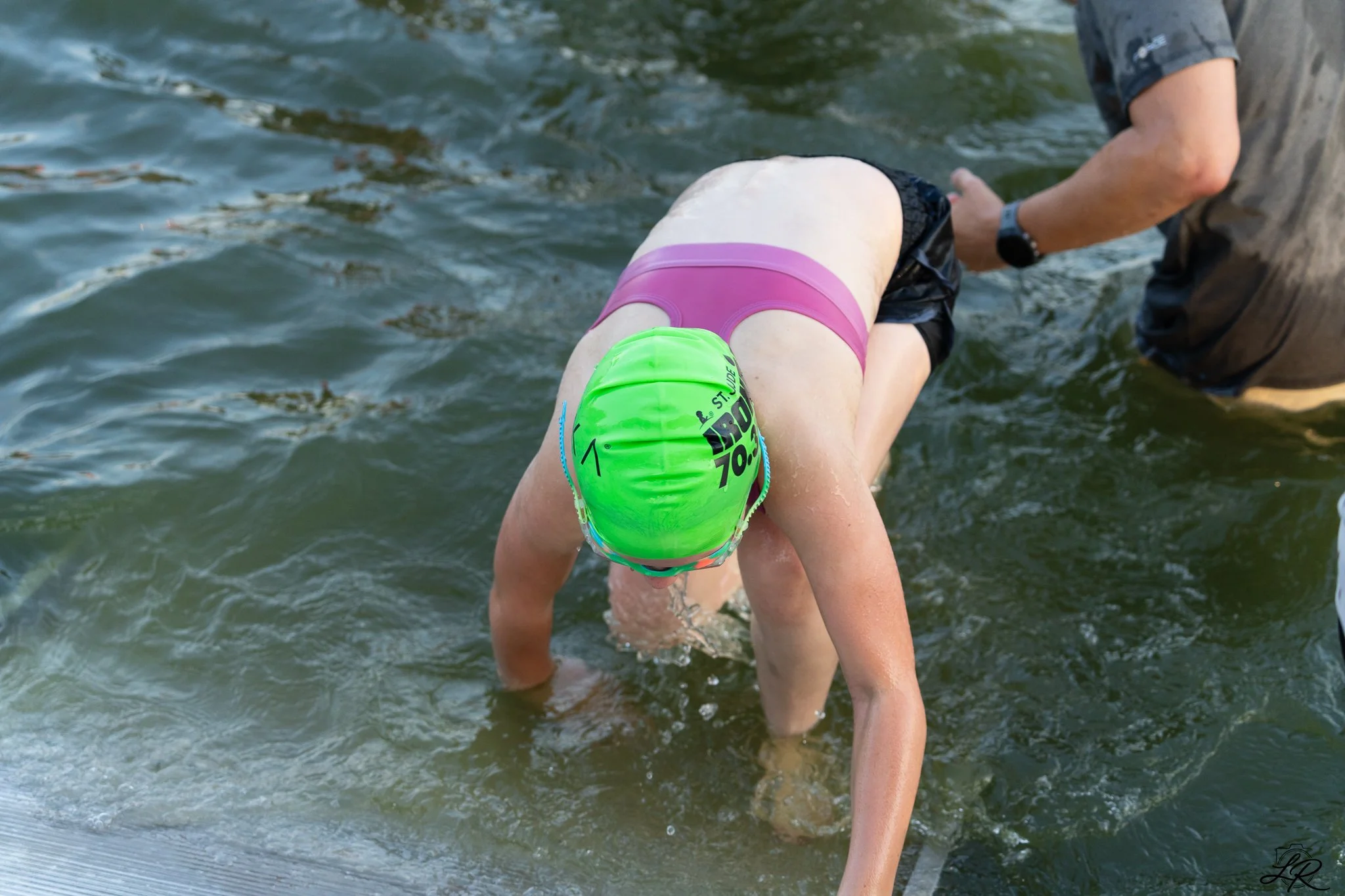 A person in a neon green swim cap and pink swimsuit is leaning forward in the water, with another person assisting nearby, in an outdoor aquatic setting.