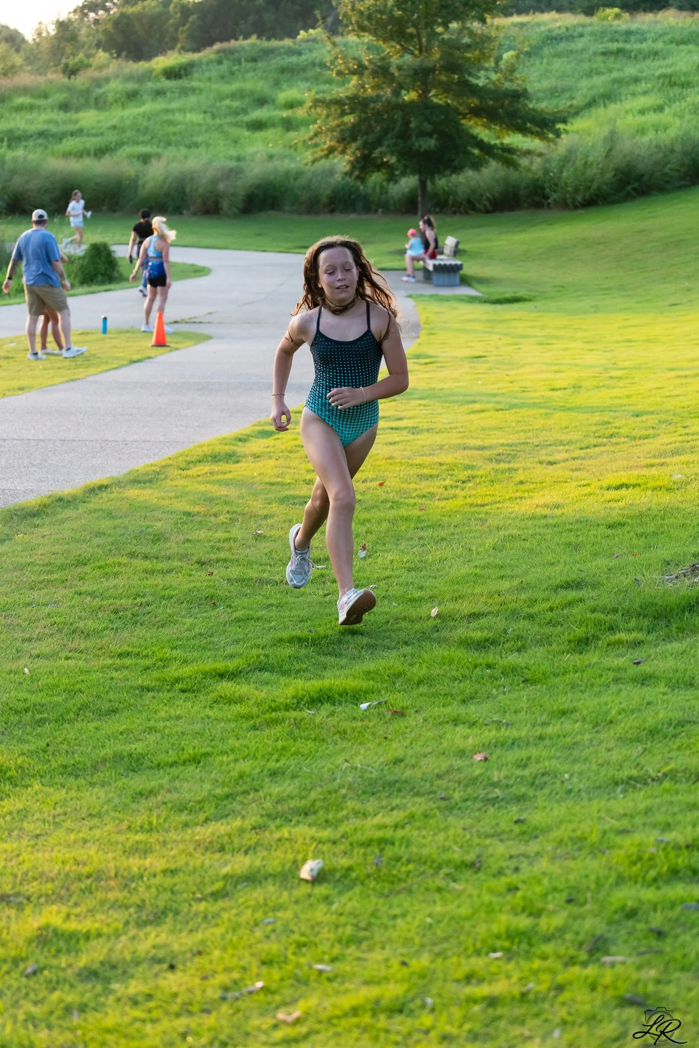 A young girl with red hair running on a grassy park area, with other people in the background enjoying the outdoor space, trees, and a walking path during daytime.