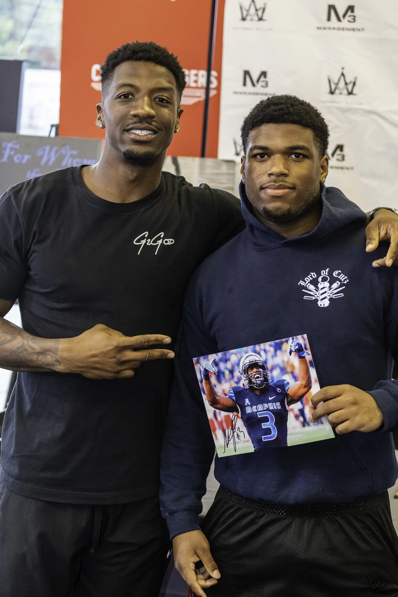 Two young men standing indoors, one wearing a black T-shirt with signature text and the other wearing a navy hoodie and holding a signed football photo of a football player in a blue Memphis jersey.