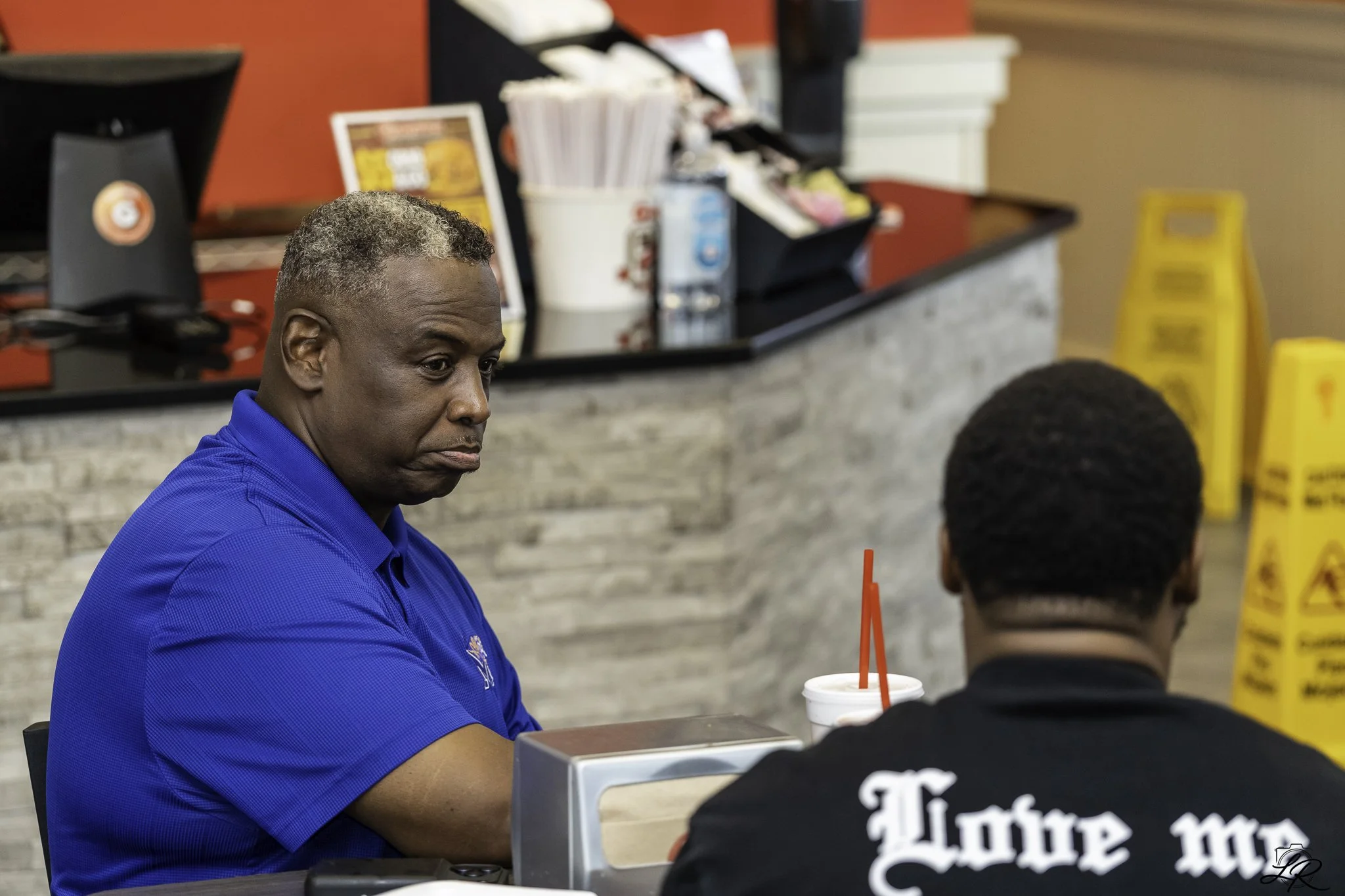 A man with gray hair and a blue shirt sitting at a table in a fast food restaurant, facing a young boy with a drink straw in his mouth, with a menu and condiment station in the background.
