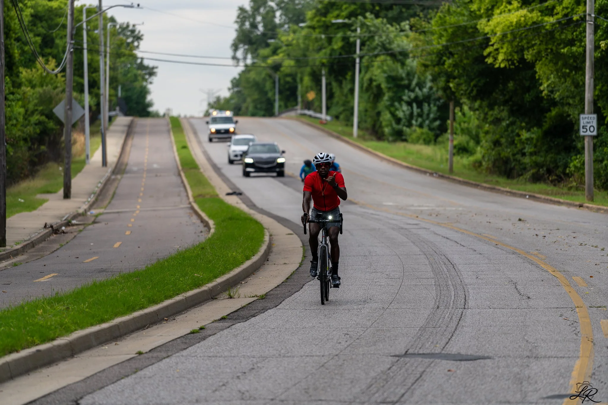 A man wearing a red shirt and helmet riding a bicycle on a suburban road with cars and two pedestrians in the background, greenery and power lines on either side, and a speed limit sign of 50 mph.