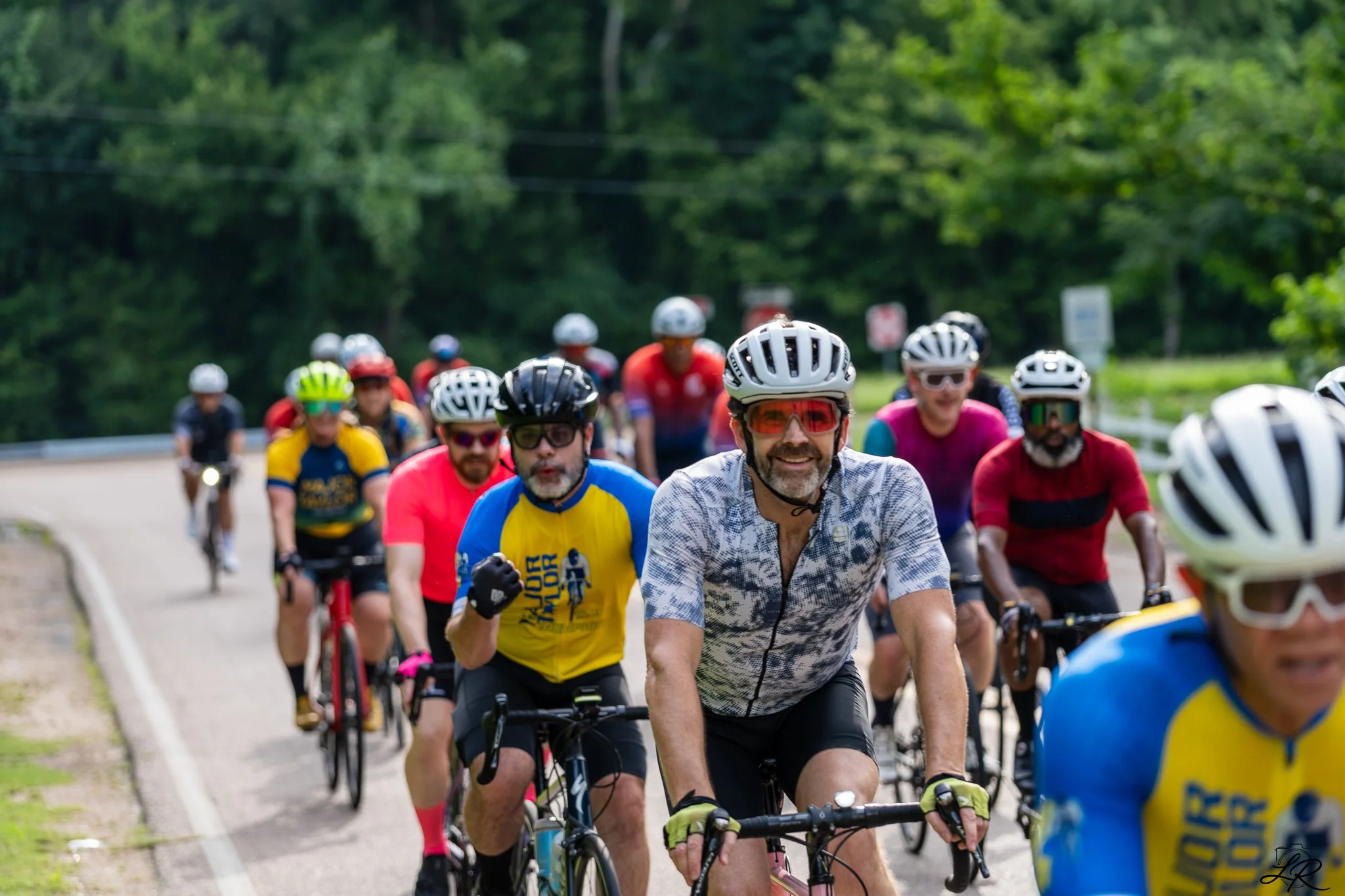 A group of cyclists riding on a road surrounded by green trees, led by a man with sunglasses and a patterned shirt.