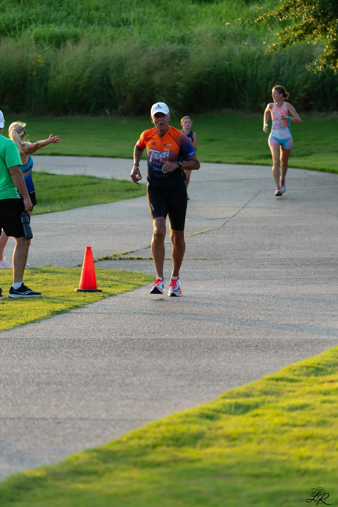 An older man running on a paved trail during a race, wearing a bright orange race shirt with logos, black shorts, a white cap, and white running shoes. A woman in colorful workout clothes and pink running shoes runs behind him. A woman in a green shi