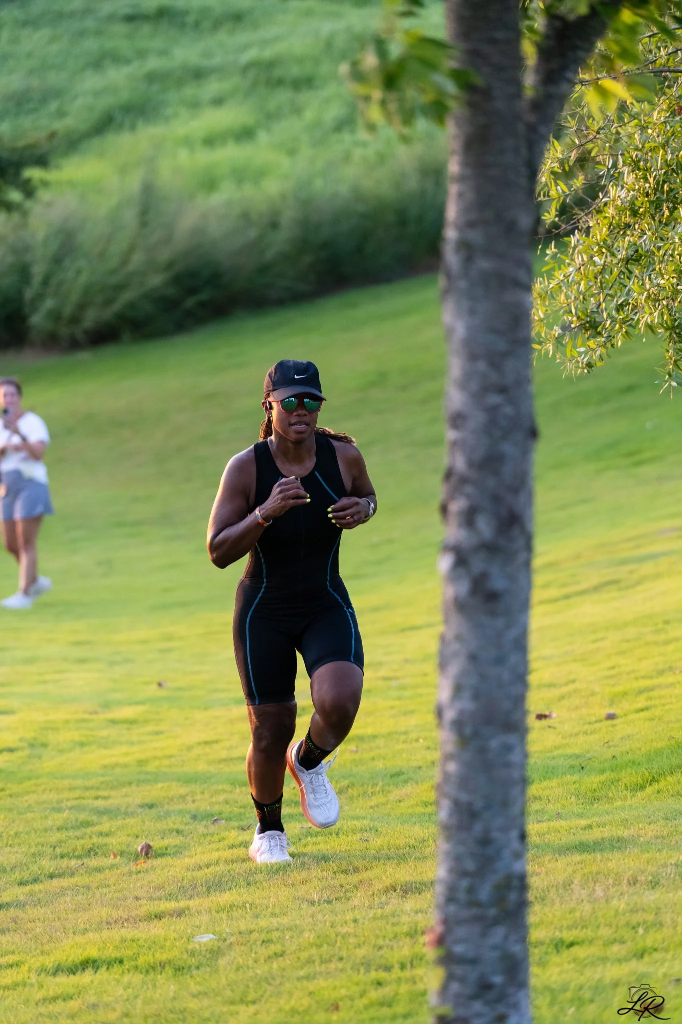A woman running in a park, wearing black athletic gear, a black cap, sunglasses, and white sneakers, with trees and grass in the background.
