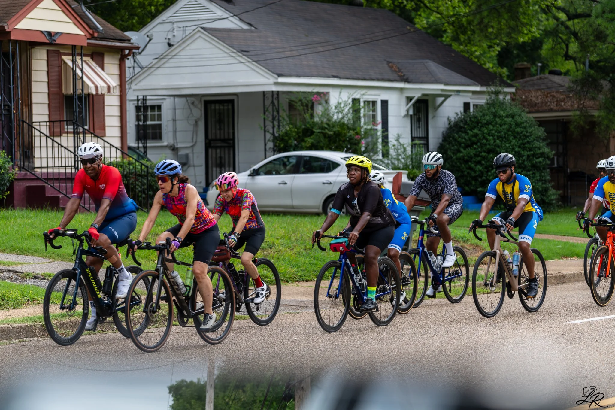 Group of people riding bikes on a neighborhood street with houses and trees in the background.