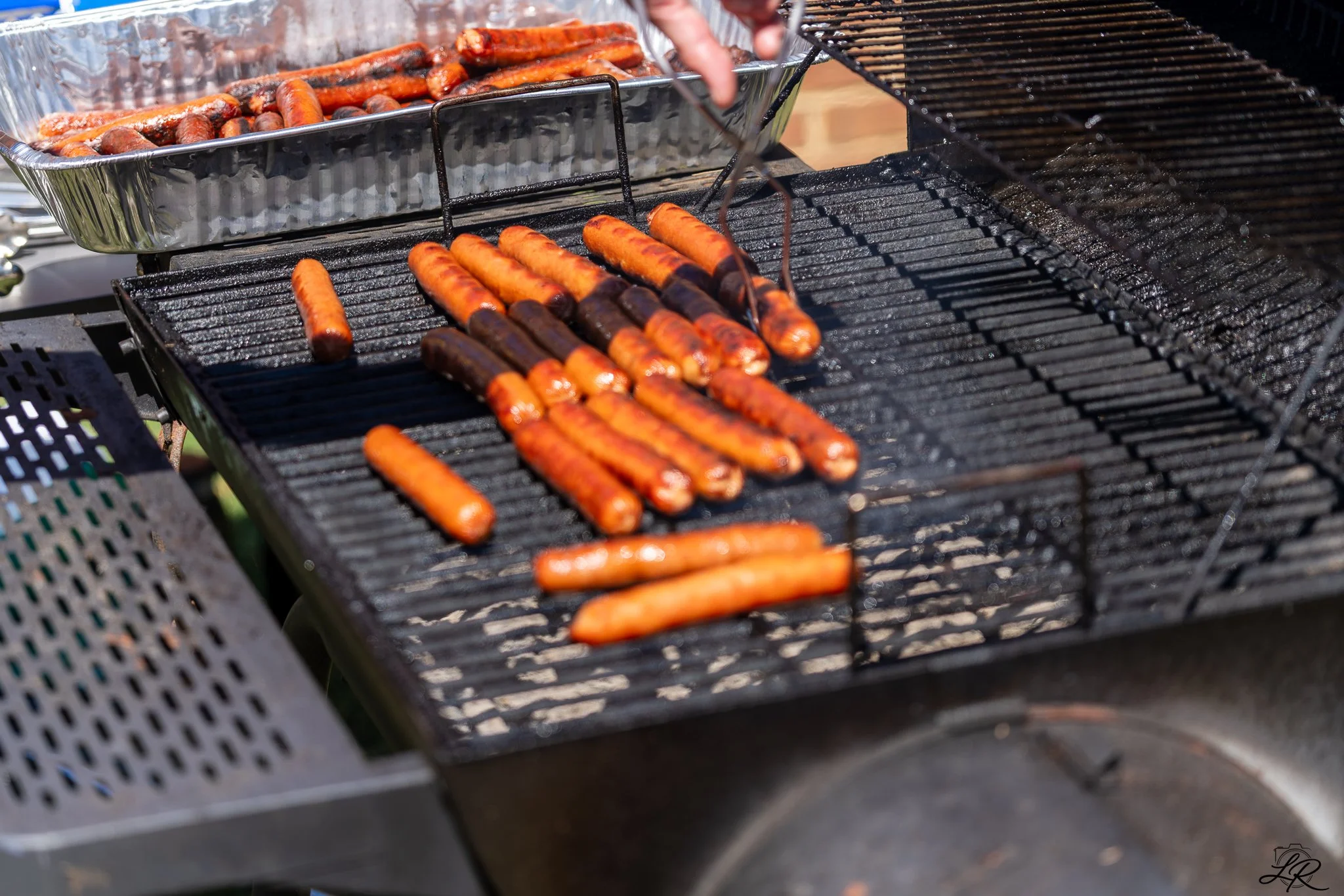 Sausages being grilled on a barbecue with a tray of more cooked sausages in the background.