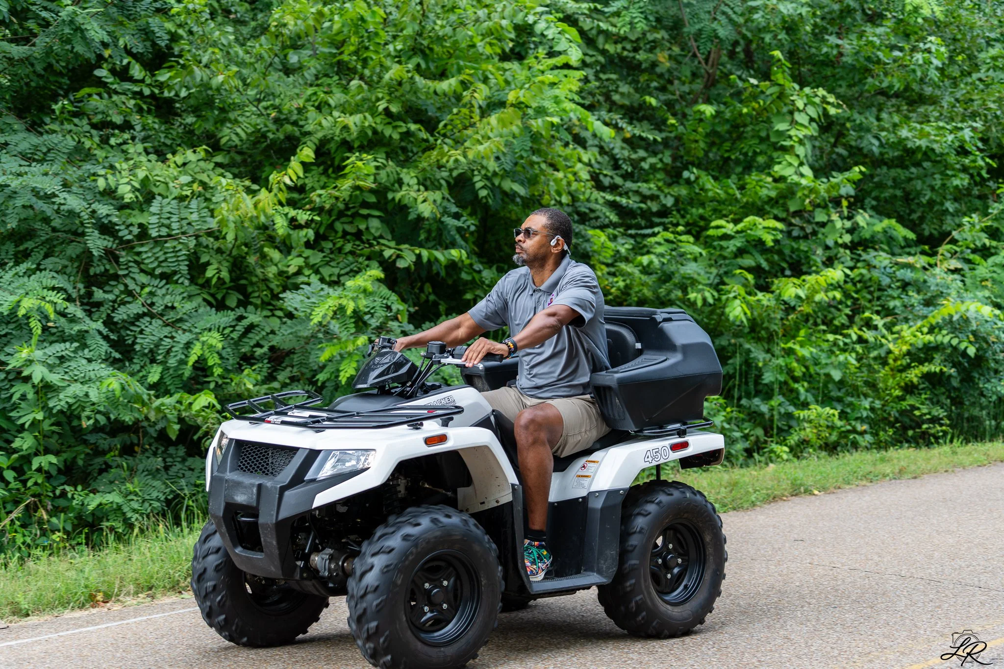A man riding an all-terrain vehicle (ATV) on a dirt trail by dense green foliage, wearing sunglasses, a gray polo shirt, tan shorts, and sneakers.