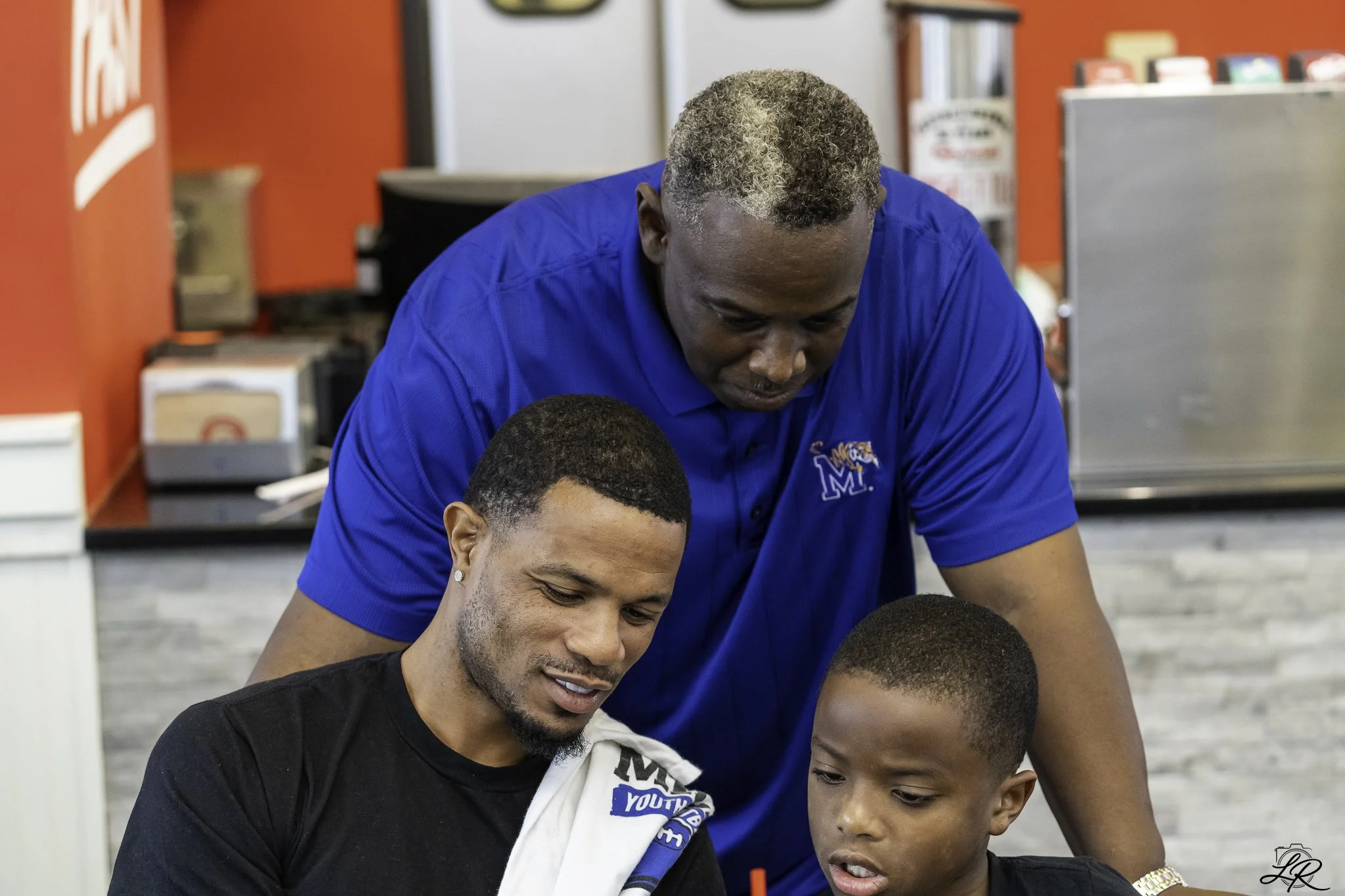 A man with gray and black curly hair wearing a blue polo shirt leaning over and looking at two young boys, one with short dark hair and a black shirt, and the other with a shaved head and a black shirt, inside a room with red walls.