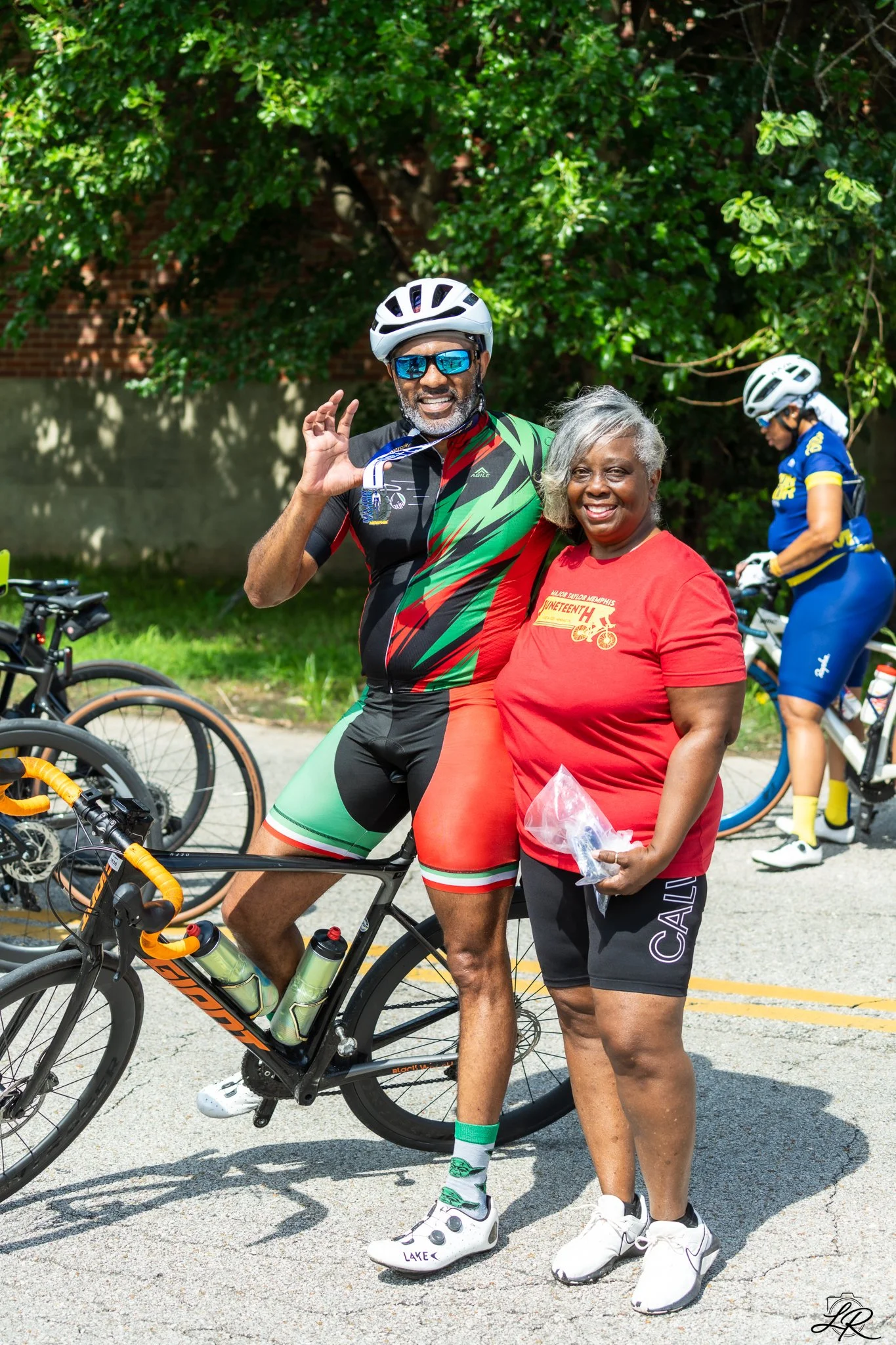 A man and an older woman standing together outdoors, smiling for a photo. The man is wearing cycling gear, including a helmet, sunglasses, and a colorful cycling outfit, with his bike visible. The woman is wearing a red t-shirt and black shorts, hold