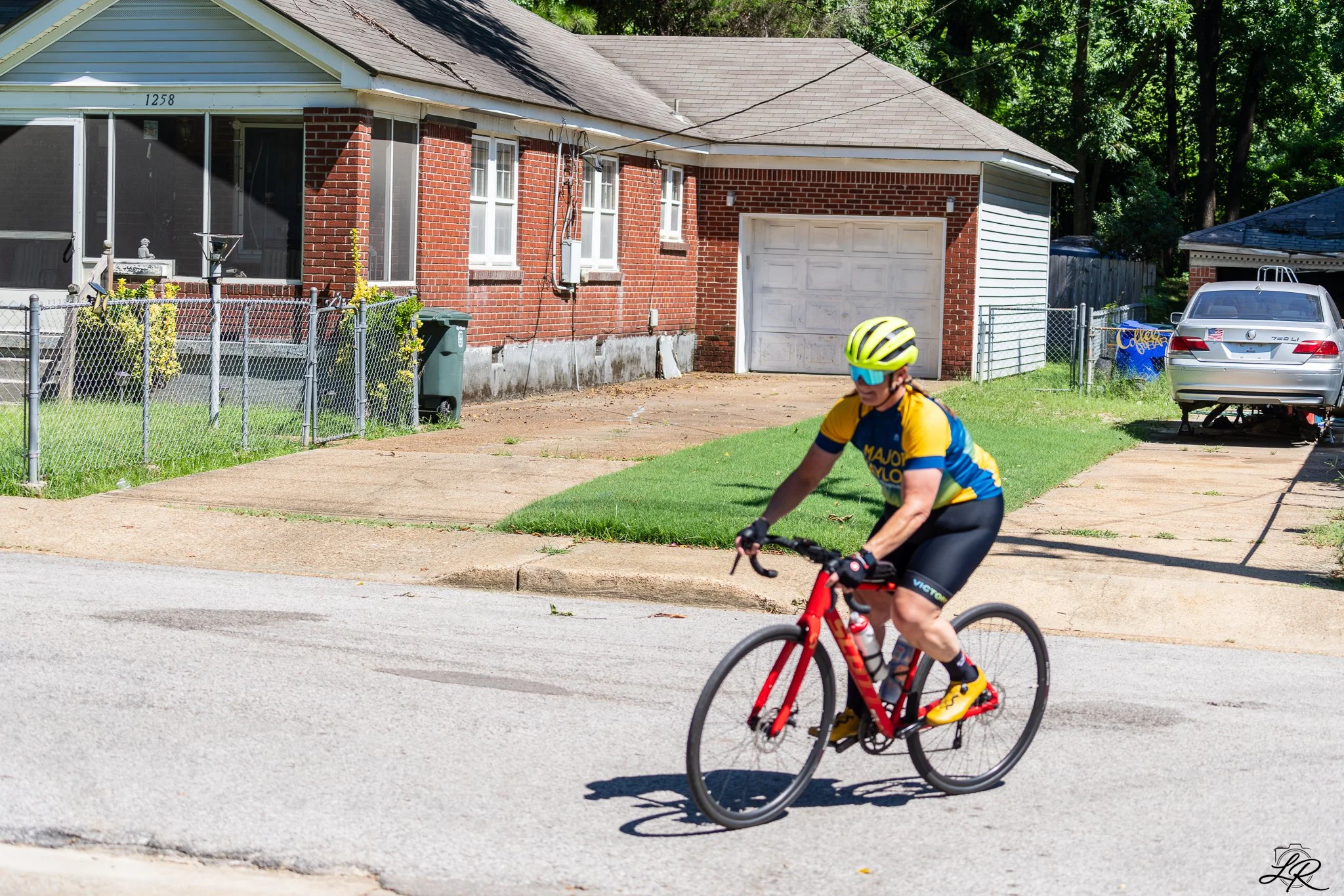 A young male cyclist wearing a yellow and blue cycling jersey, black shorts, sunglasses, and a yellow helmet riding a red bicycle on a residential street. In the background, there is a brick house with a white garage door, a fenced yard, a green tras