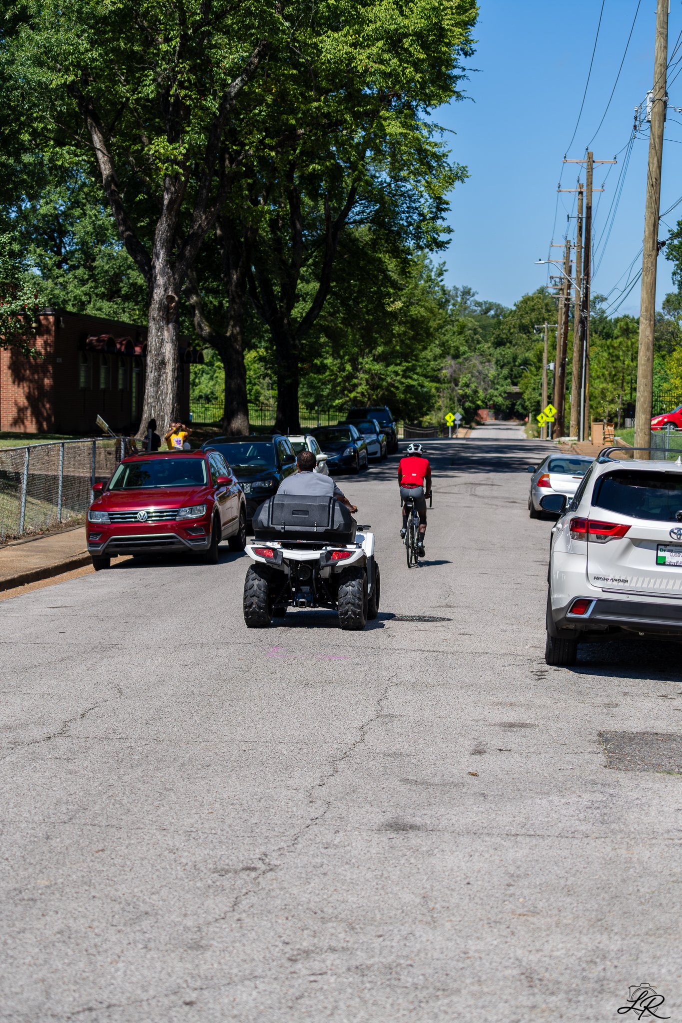 A street scene with cars parked along the side of the road, a person riding a bicycle, and trees lining the street under a blue sky.