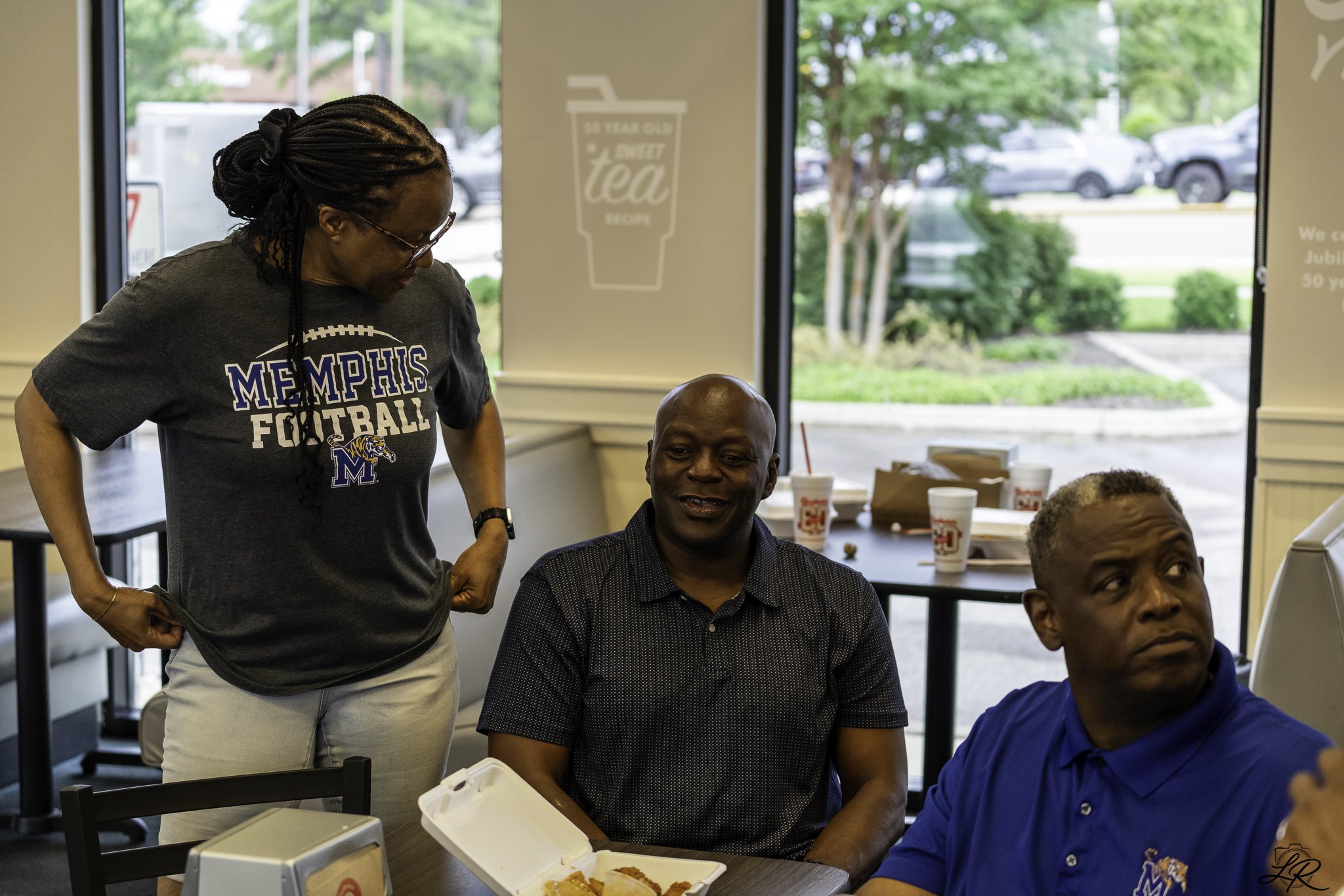 Three men seated at a table inside a fast-food restaurant with one woman standing nearby. The woman is wearing a Memphis football t-shirt and glasses, and is leaning toward the men. The table has takeout food containers and cups. Large windows show c