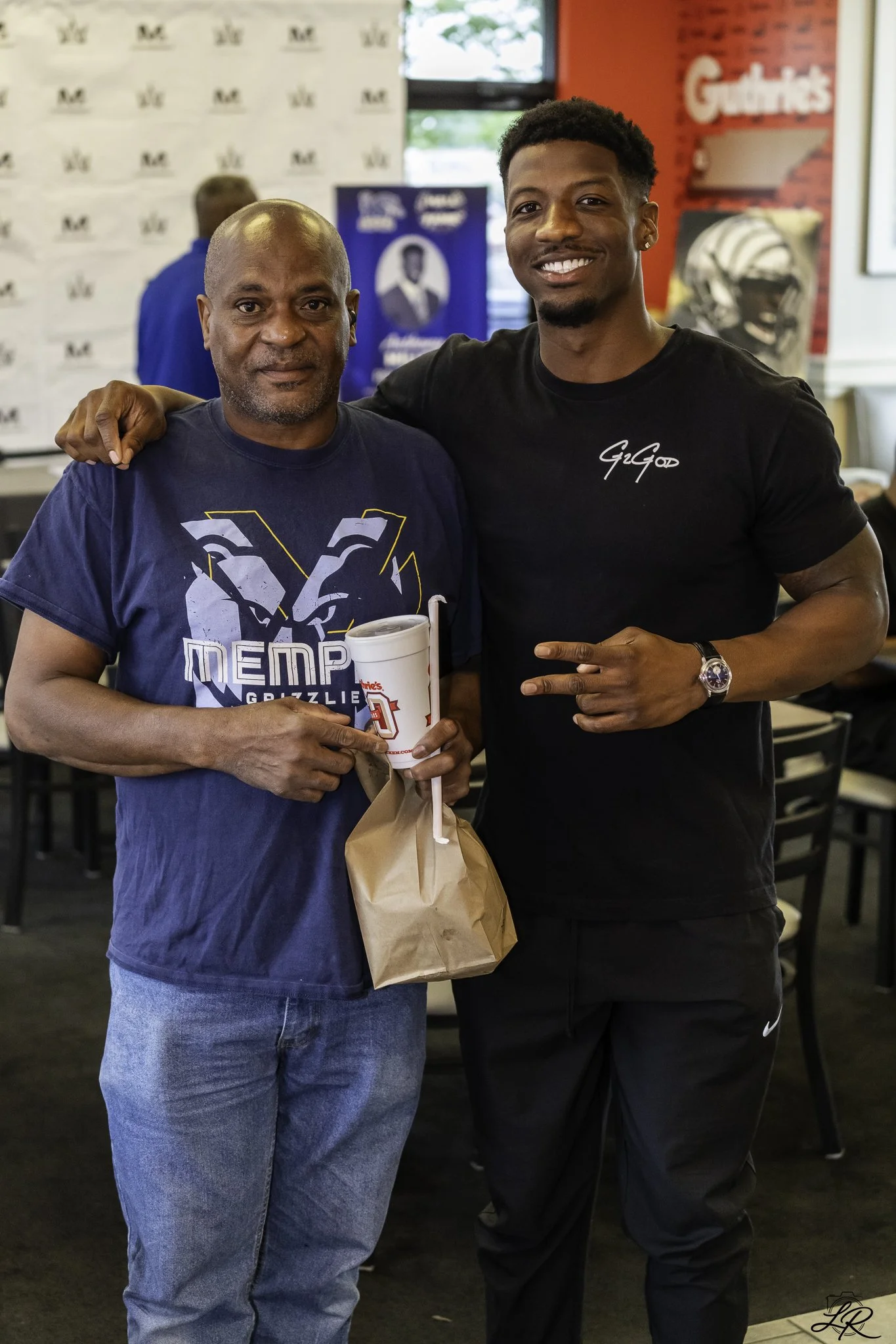Two men standing together inside a restaurant or fast food place, smiling at the camera. The man on the left is wearing a blue Memphis Grizzlies t-shirt and holding a fast food drink in a cup. The man on the right is wearing a black t-shirt with whit