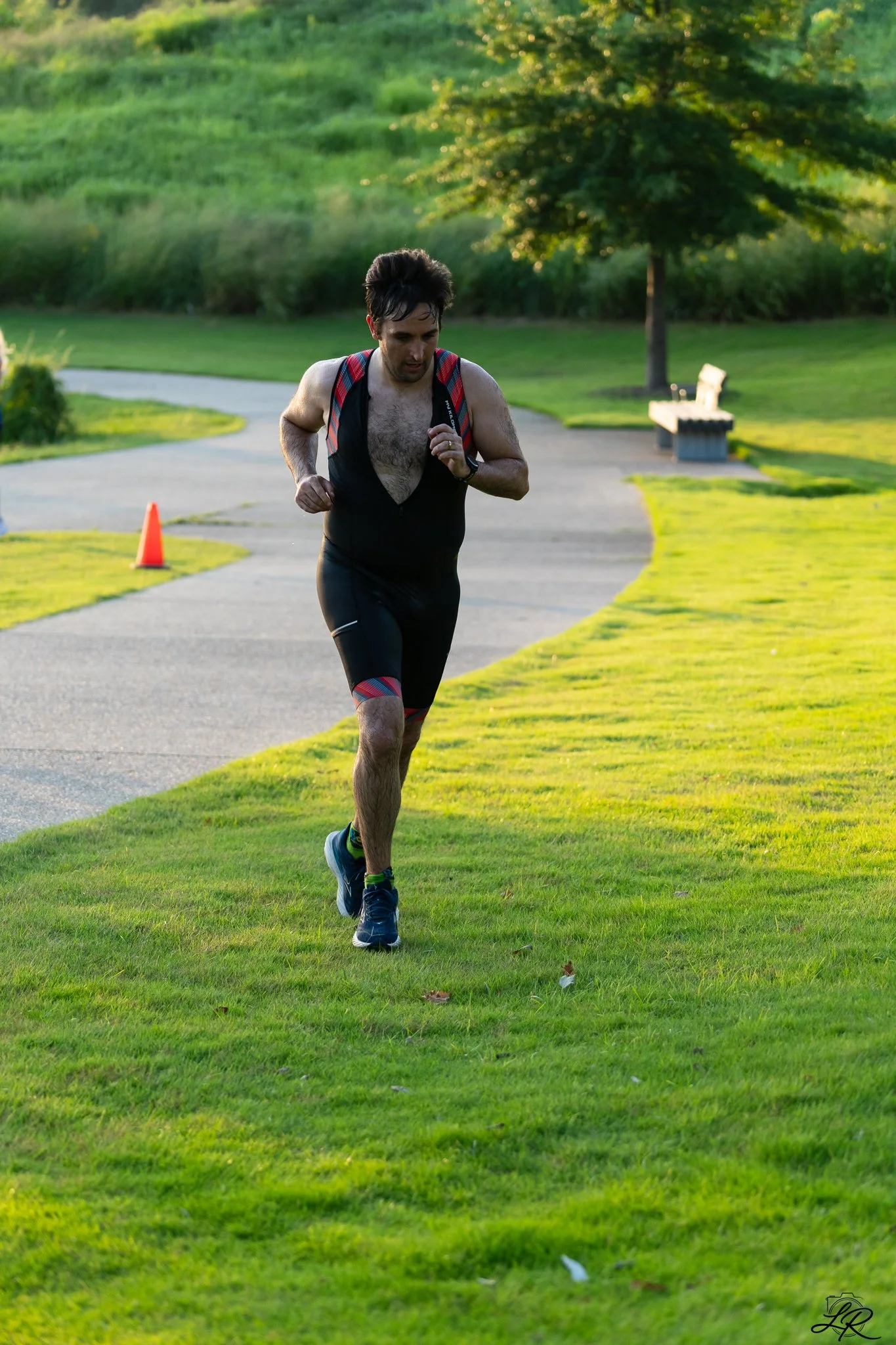 A man running on a grassy park trail during sunset, wearing athletic clothing and a backpack.