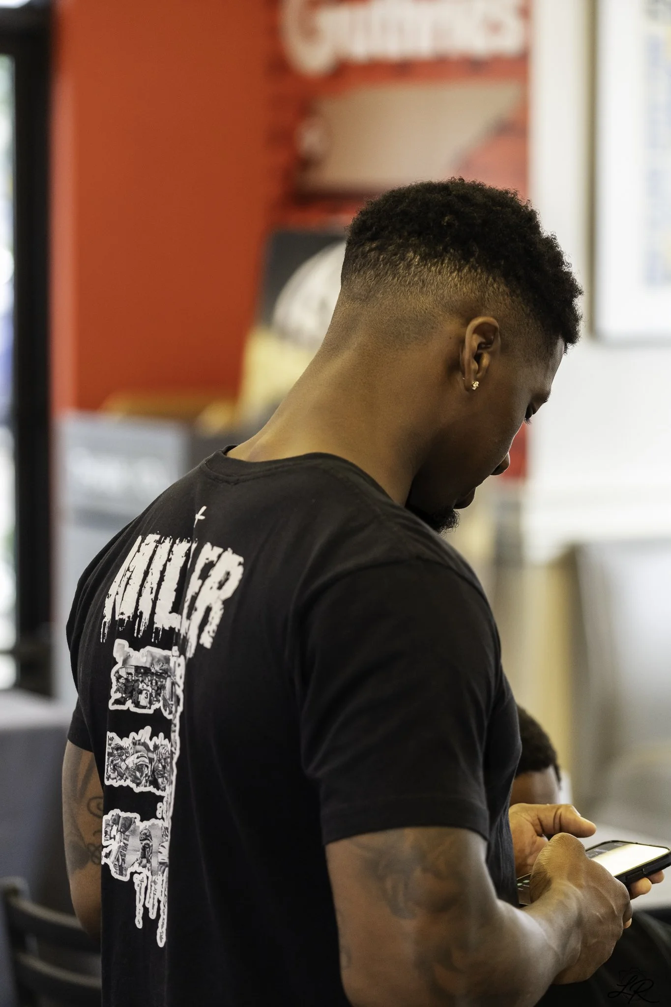 A man with short curly hair and earrings, wearing a black graphic T-shirt, is looking at and using a smartphone with both hands. The background is blurred but shows a red wall with a partial advertisement or sign.