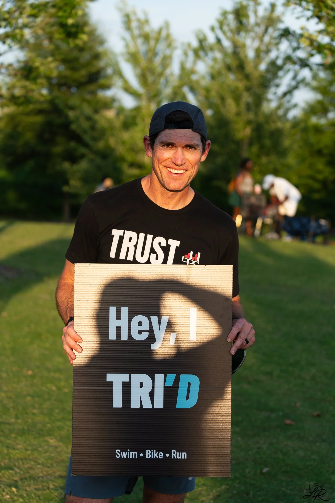 Man smiling, wearing a black shirt with the word 'TRUST' and a wearable device graphic, holding a sign that says 'Hey, I TR' with the rest covered, standing outdoors in a park with other people in the background.