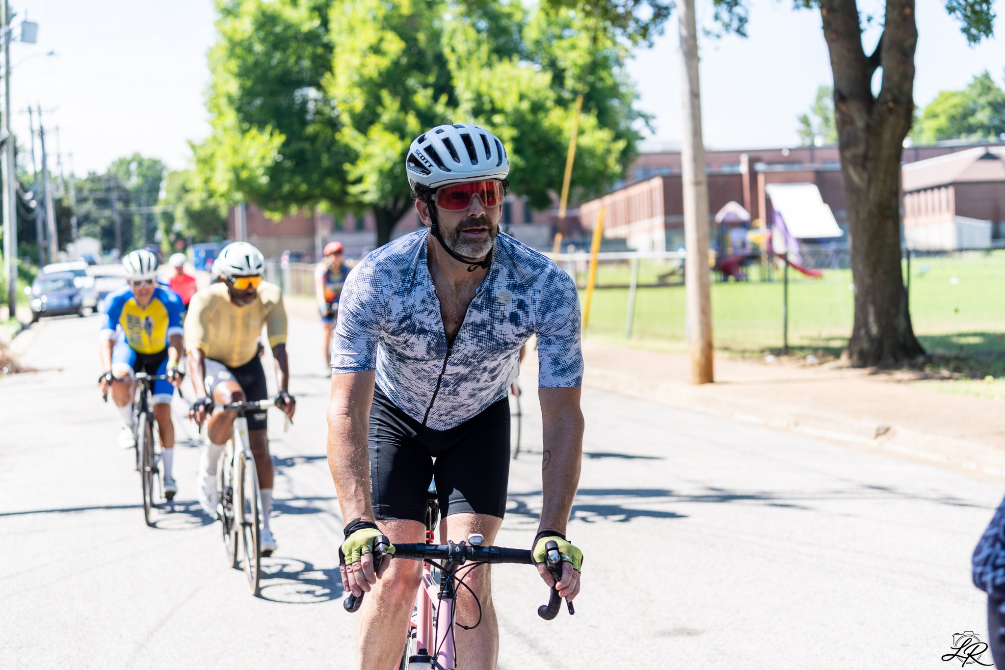 A group of cyclists riding on a sunny street lined with trees and buildings, with a man in the front wearing a helmet and sunglasses.