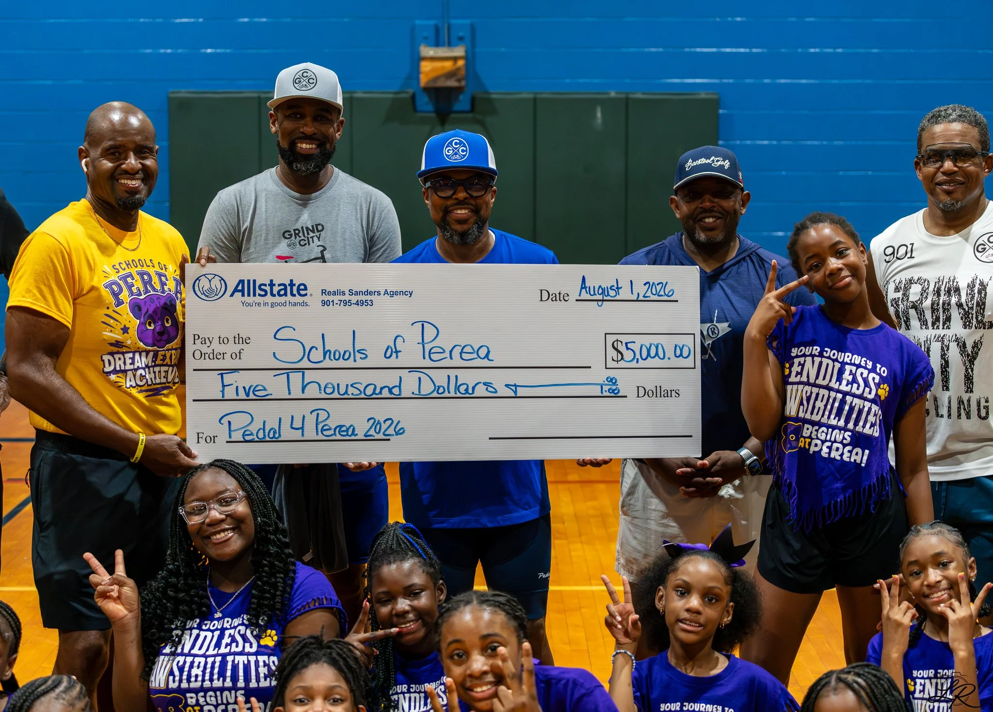 Group of smiling individuals, including children and adults, holding a large check for $5,000 made out to Schools of Perea, dated August 1, 2026, inside a gymnasium gym.