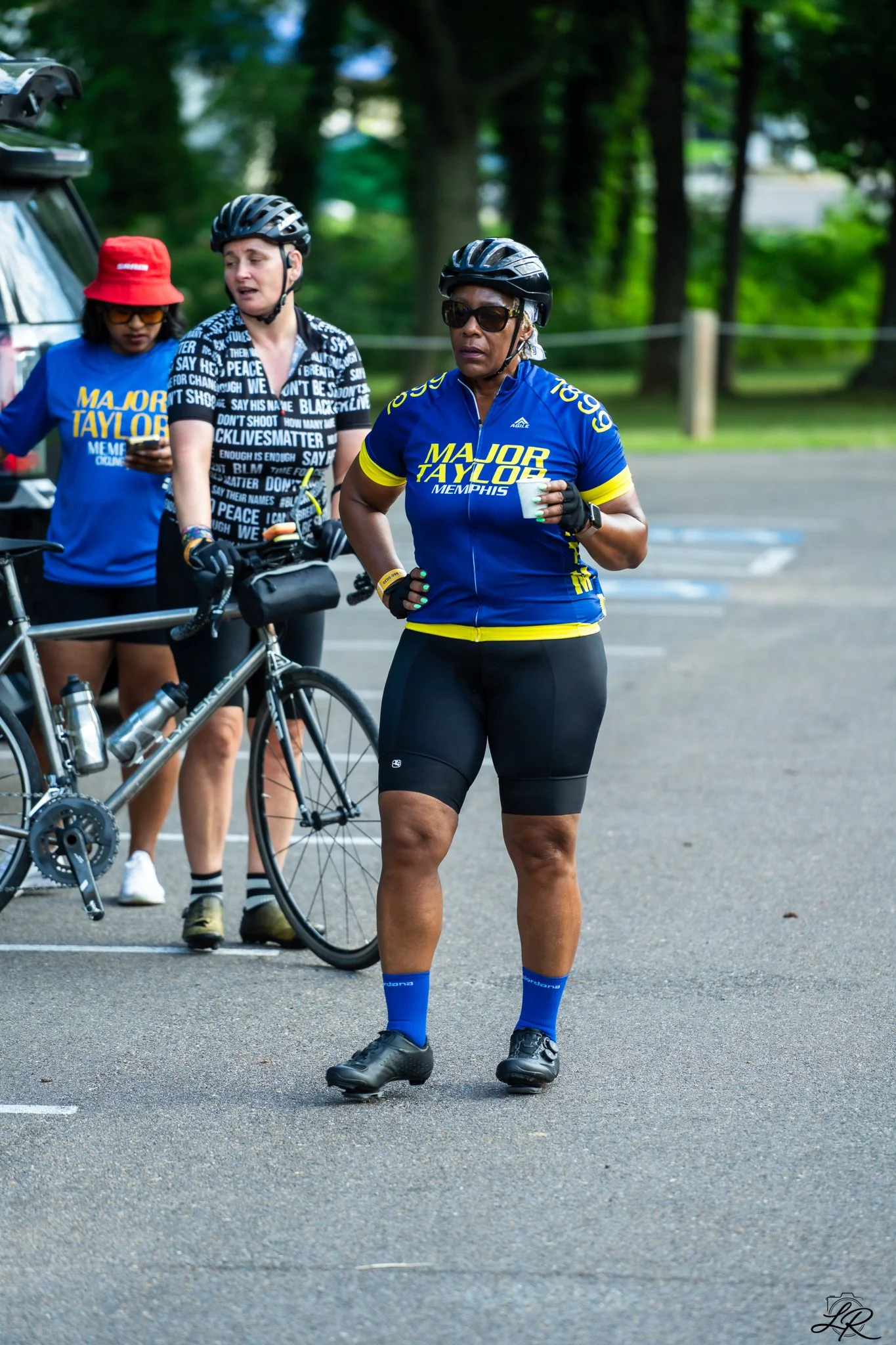 Three women, two of whom are wearing black helmets, black cycling shorts, and jerseys with 'Major Taylor Memphis' written on them. One woman is holding a bicycle, while the other two are standing, with one holding a disposable cup. The background sho