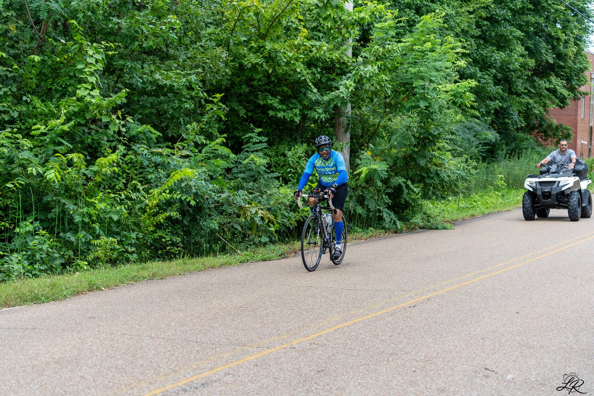 A man riding a bicycle on a paved road next to dense green foliage, with another man riding a four-wheeler behind him, in an outdoor setting.