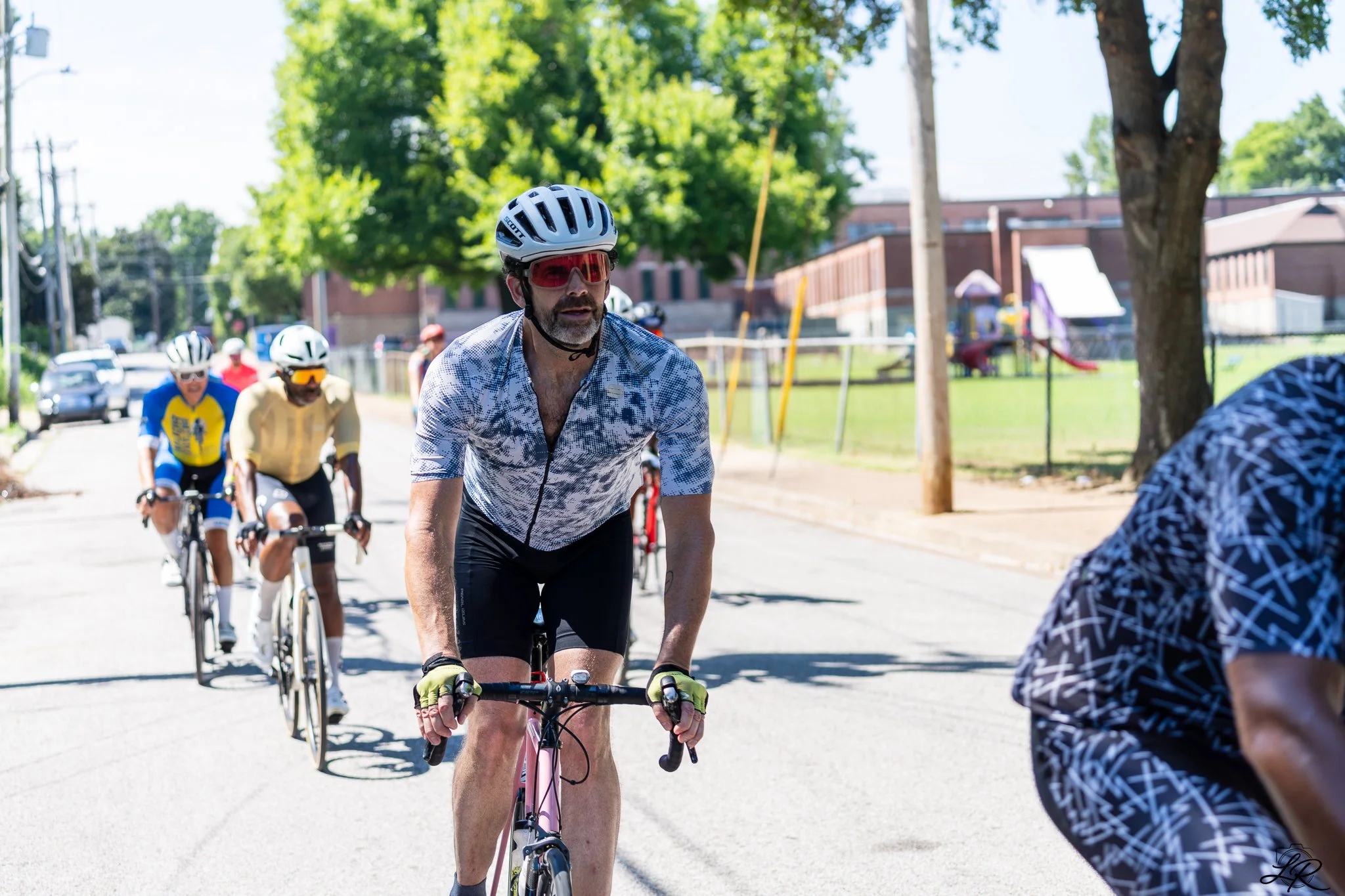 A group of people riding bicycles outdoors on a sunny day, with a man in the foreground wearing a helmet and sunglasses, lookin' ahead. There are trees, a playground, and some cars in the background.