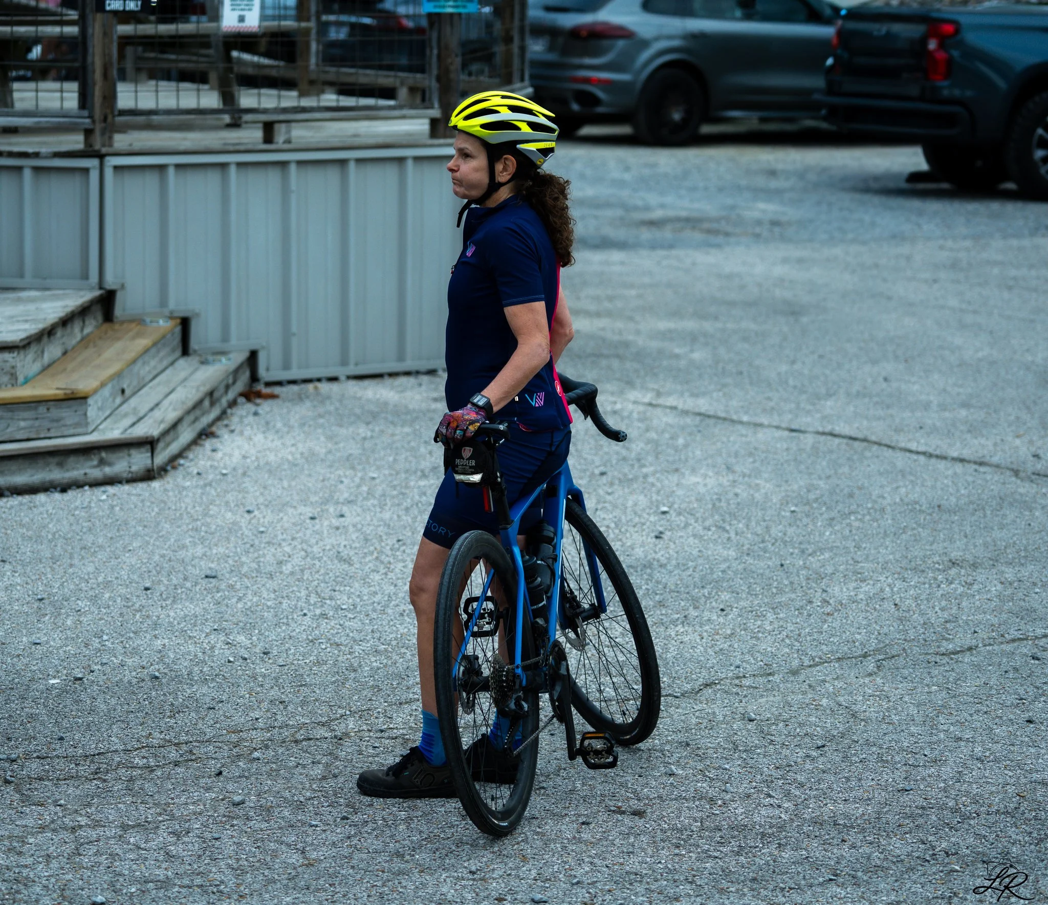 A woman in cycling gear, including a yellow helmet and blue athletic attire, standing with a blue bicycle on a gravel surface near parked cars and a metal fence.