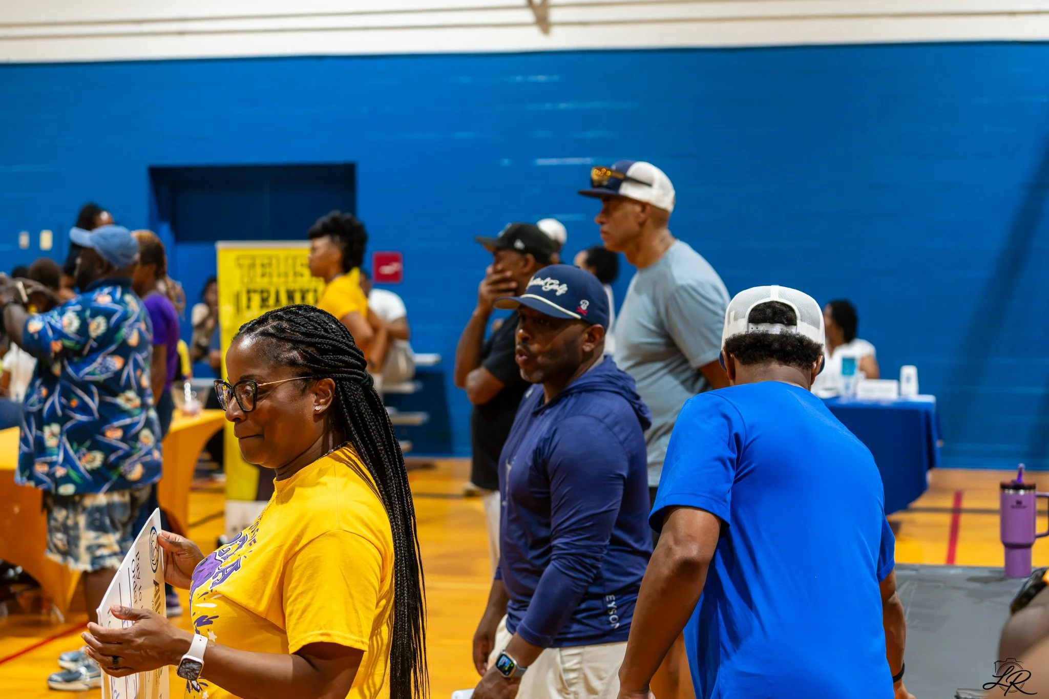 A group of people standing in line inside a gymnasium, some wearing hats and glasses, with yellow and blue clothing, facing towards the left side of the image. The background has a blue wall and a yellow sign.