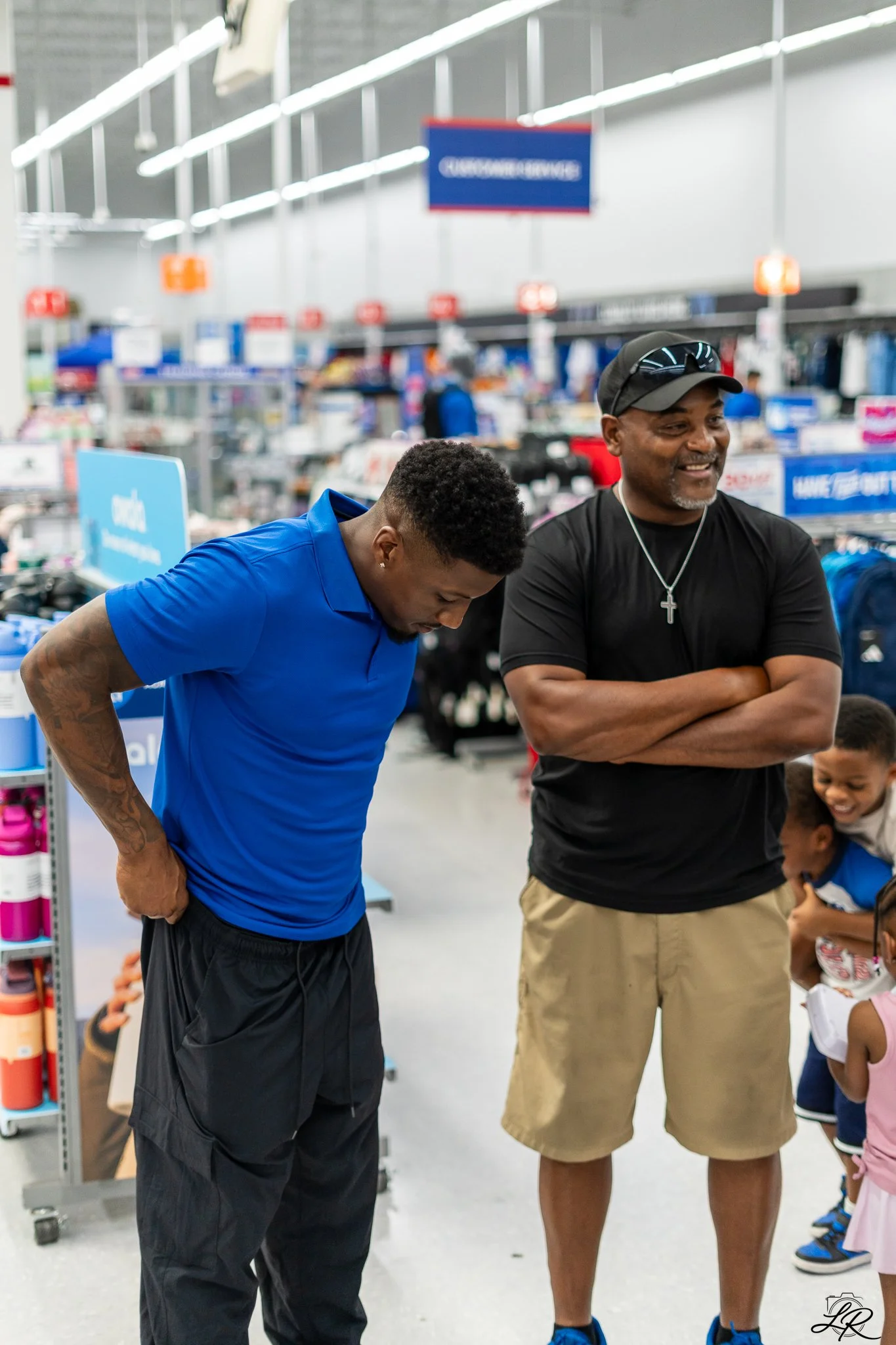 Two men and a group of children inside a retail store, with shelves and signage in the background. The man on the left is wearing a blue shirt and black pants, looking downward. The man on the right is wearing a black shirt, tan shorts, sunglasses on