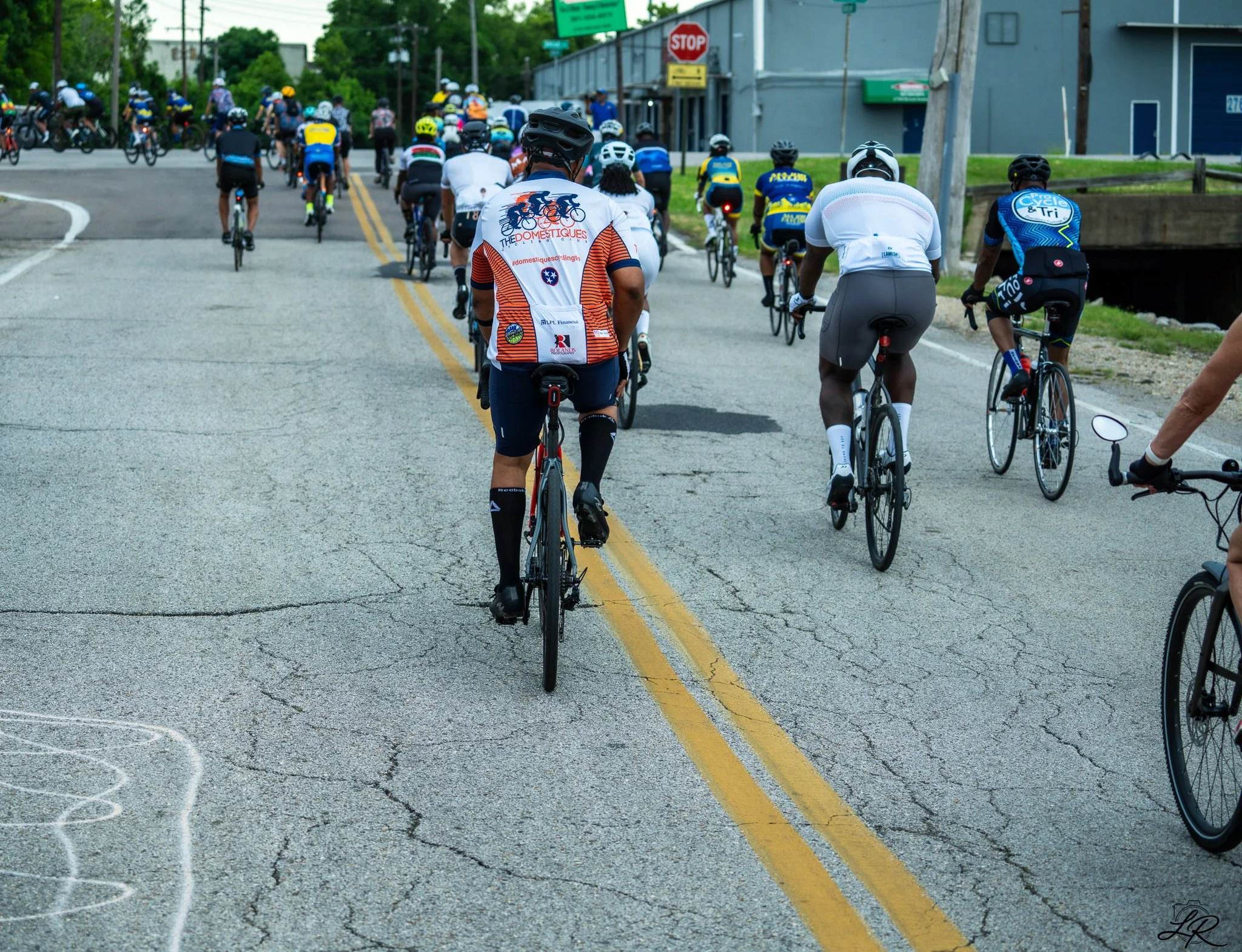 A group of cyclists riding on a city street with buildings and a stop sign in the background.