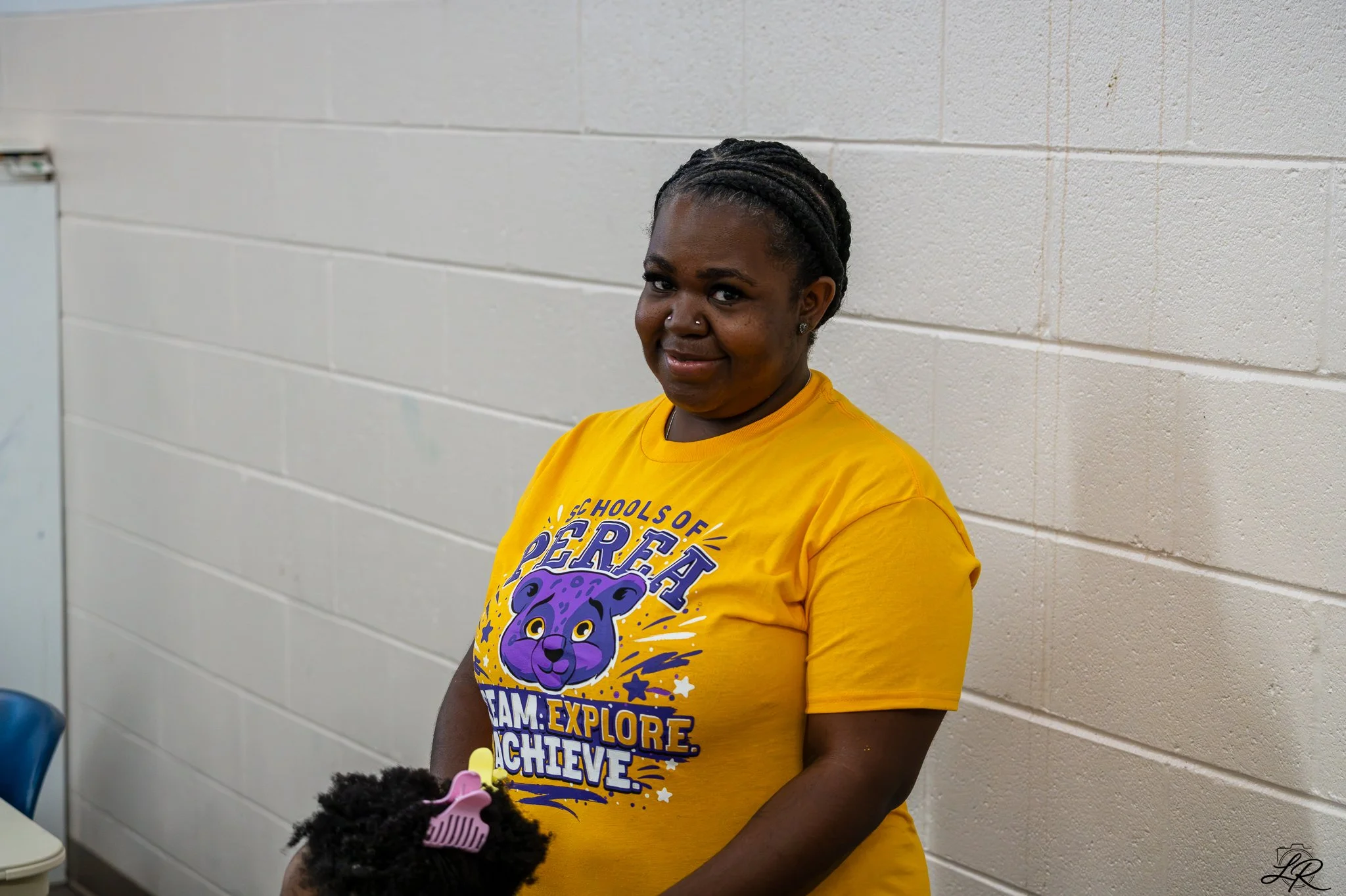 A woman with braids and a nose piercing stands against a white brick wall, wearing a yellow t-shirt with a purple panther logo and motivational text.