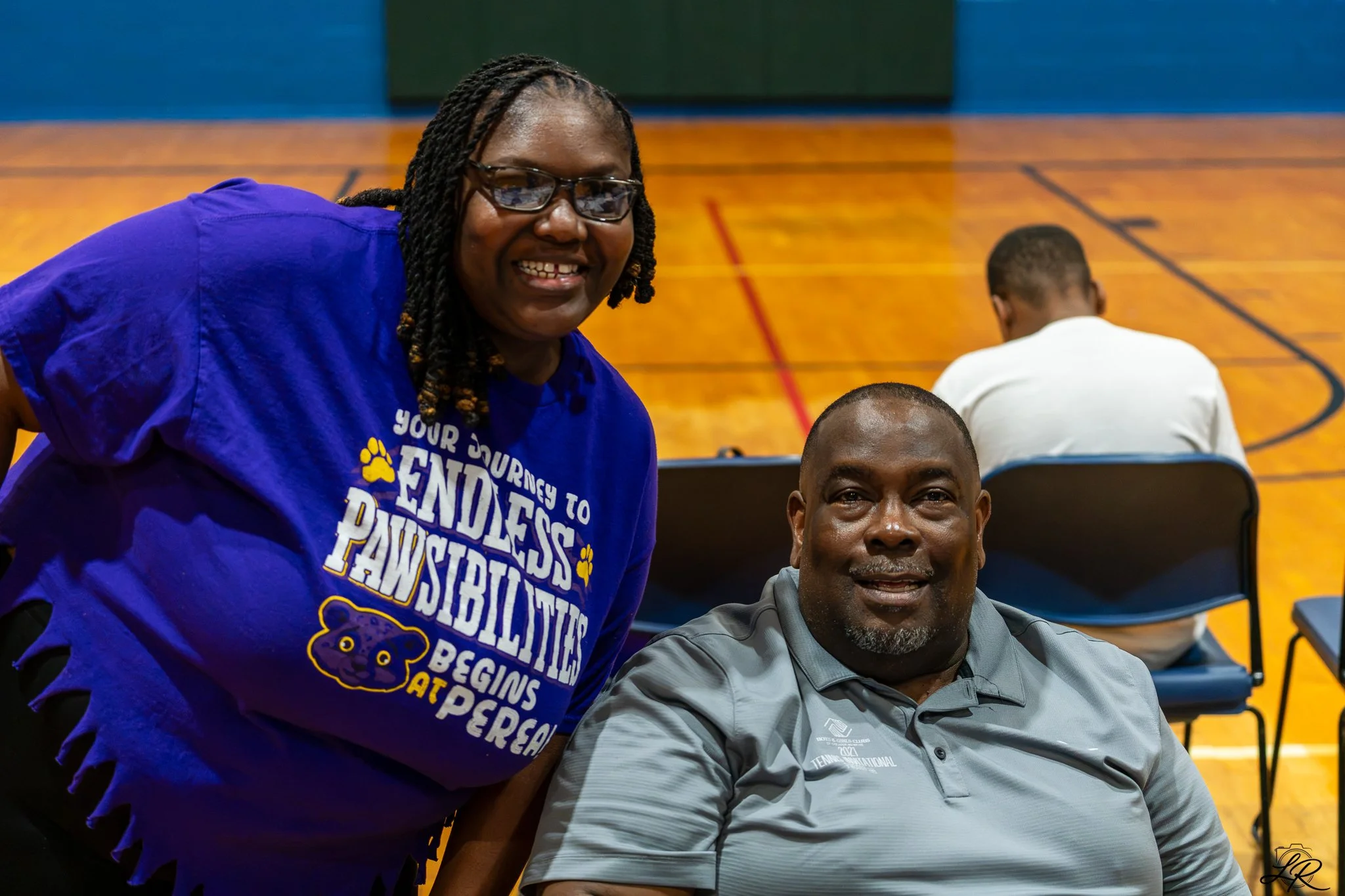 A woman with glasses and twists hairstyle wearing a blue t-shirt with yellow paw print and text, smiling at the camera, standing next to a seated man with short hair and a gray shirt, in a gymnasium.