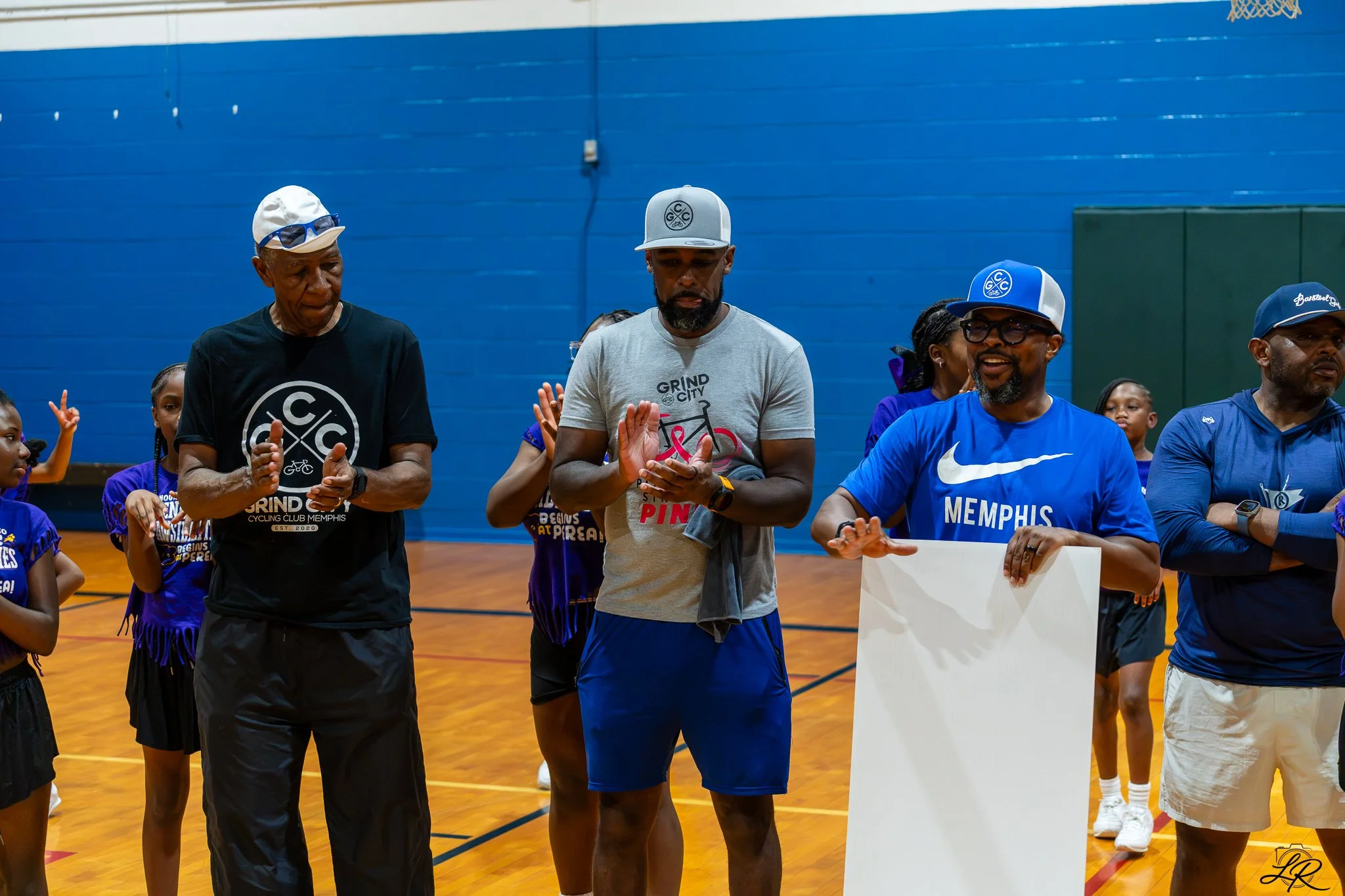 Group of African American people, including children, gathered in a gymnasium for an event. Some are wearing sportswear and caps, and appear to be praying or in reflection. The background features a blue wall and a basketball hoop.