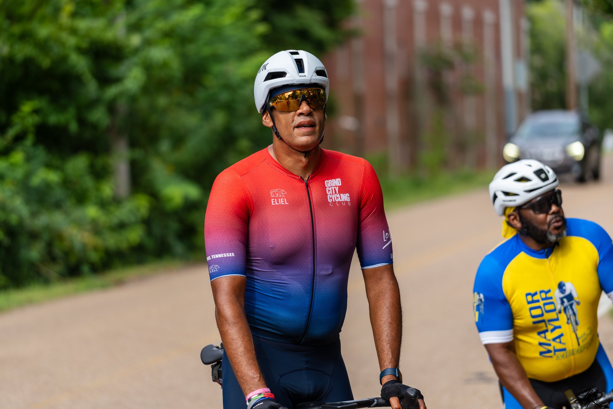 A man wearing a white bike helmet and sunglasses rides a bicycle with a man in a yellow and blue cycling jersey nearby, on a park path with green trees and a brick building in the background.