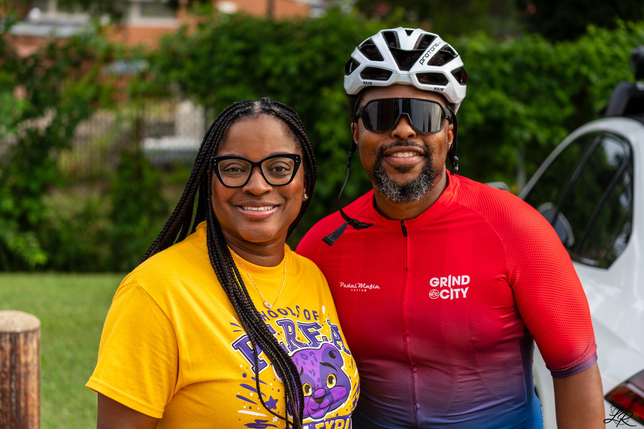 A smiling woman with glasses and braided hair standing next to a man in a bike helmet and sunglasses, both outdoors in front of green bushes.