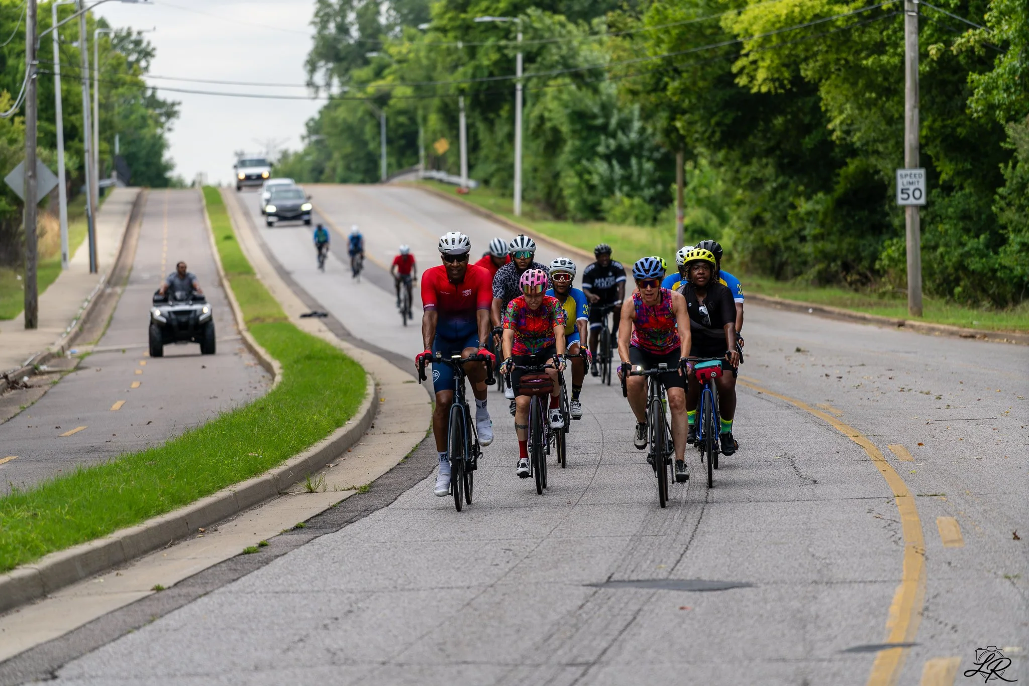 Group of cyclists riding on a road with cars and greenery on the sides.