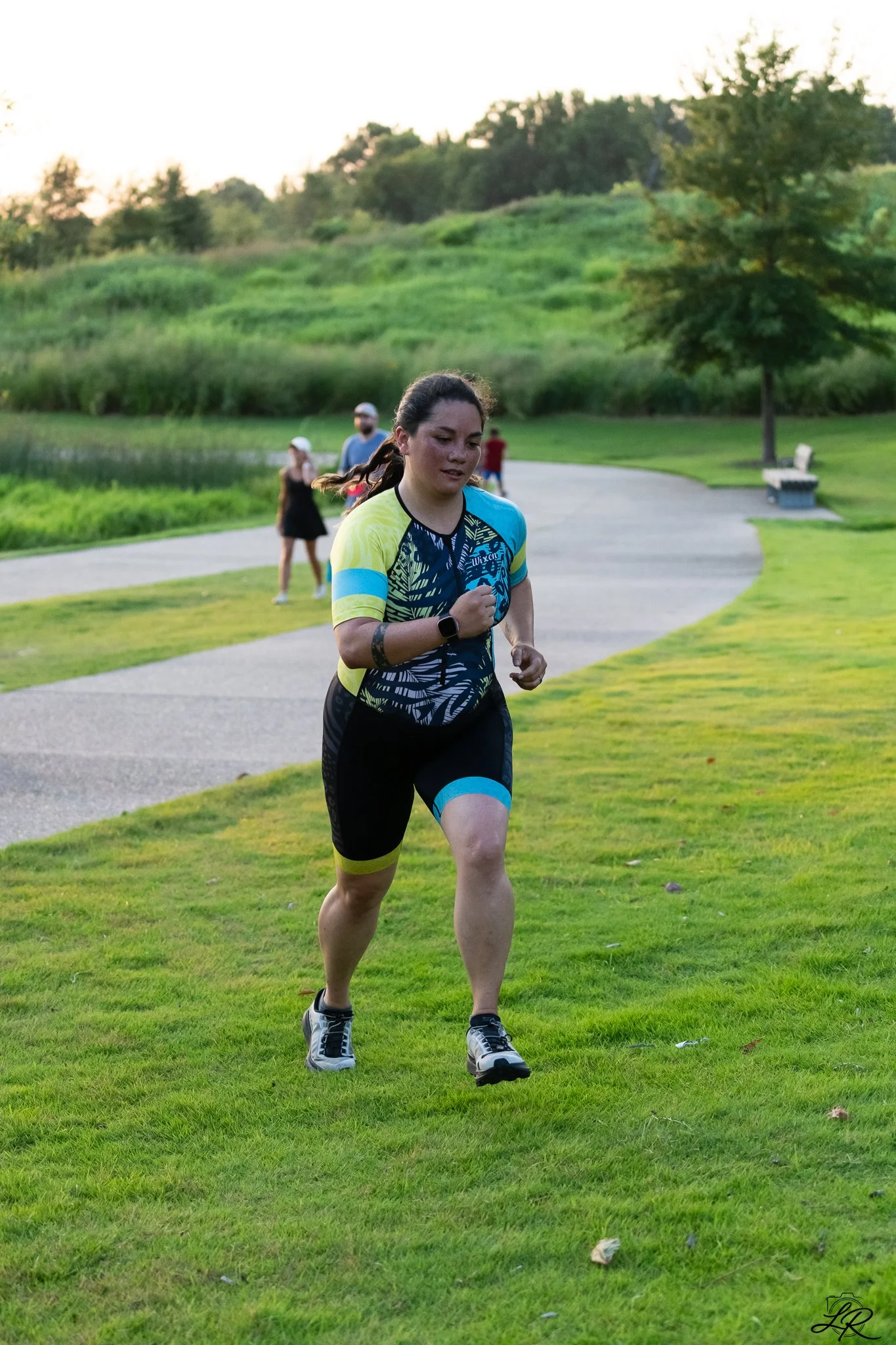 A woman running outdoors on a park path, with trees and grassy hills in the background and a group of people walking further behind her.