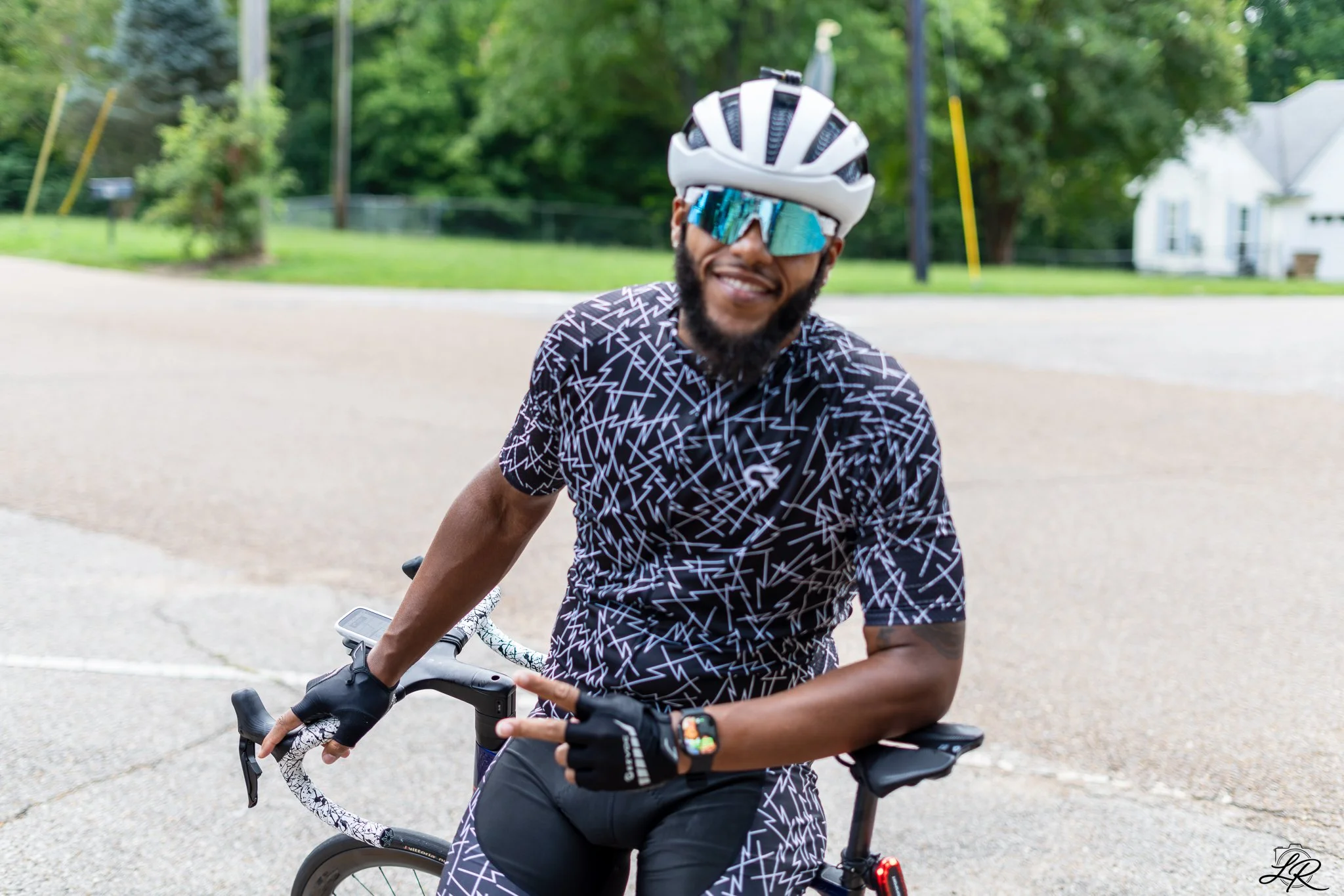 Smiling man with a beard wearing a white bike helmet, blue sunglasses, a black and white patterned cycling shirt, and gloves, sitting on a bicycle outdoors in a park with green trees and a house in the background.