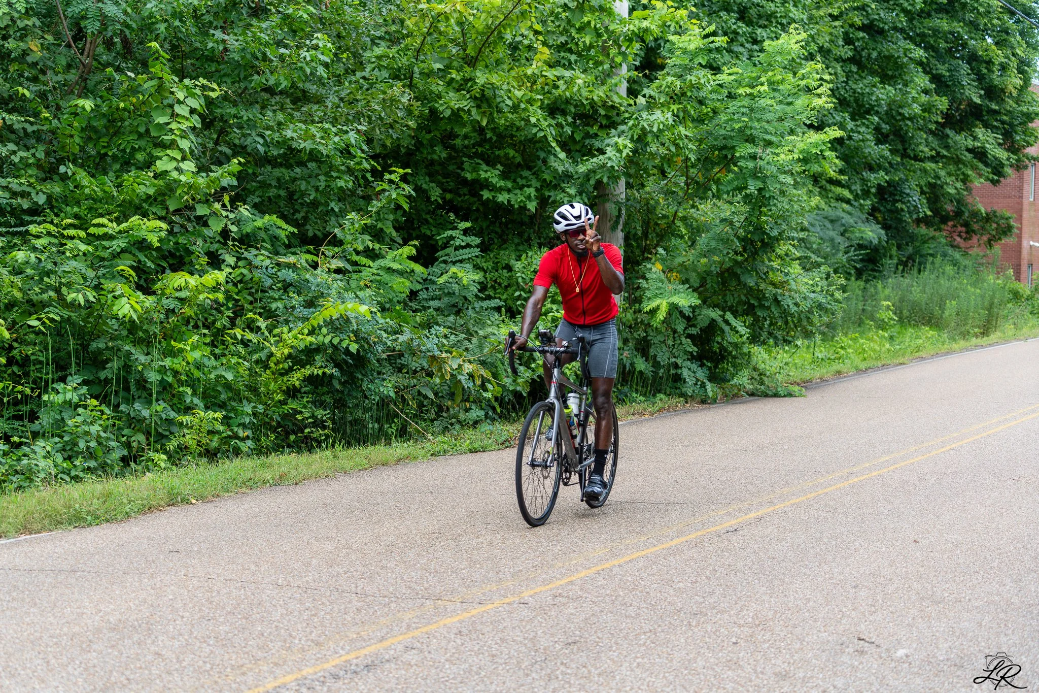 A man wearing a red shirt, sunglasses, and a white helmet riding a bicycle on a paved path next to a green bushy area with trees and a brick building in the background.