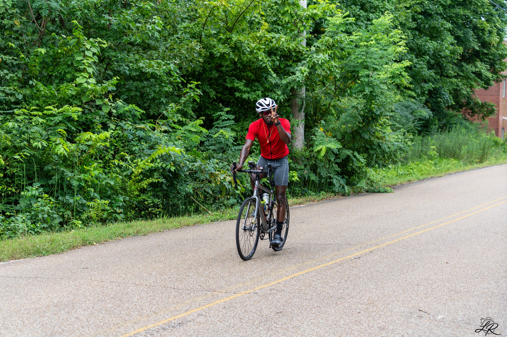A man wearing a red shirt, gray shorts, a helmet, and sunglasses riding a bicycle on a paved path lined with green trees.