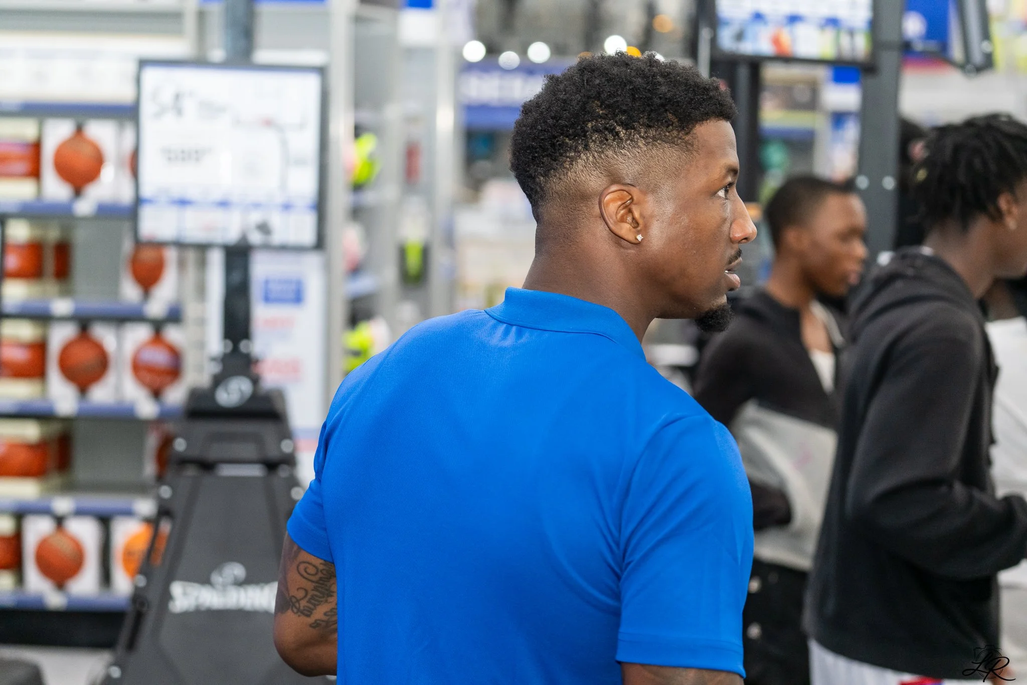 A man with short curly black hair and earrings wearing a blue shirt, sitting in a store surrounded by shelves with sports equipment, including basketballs.