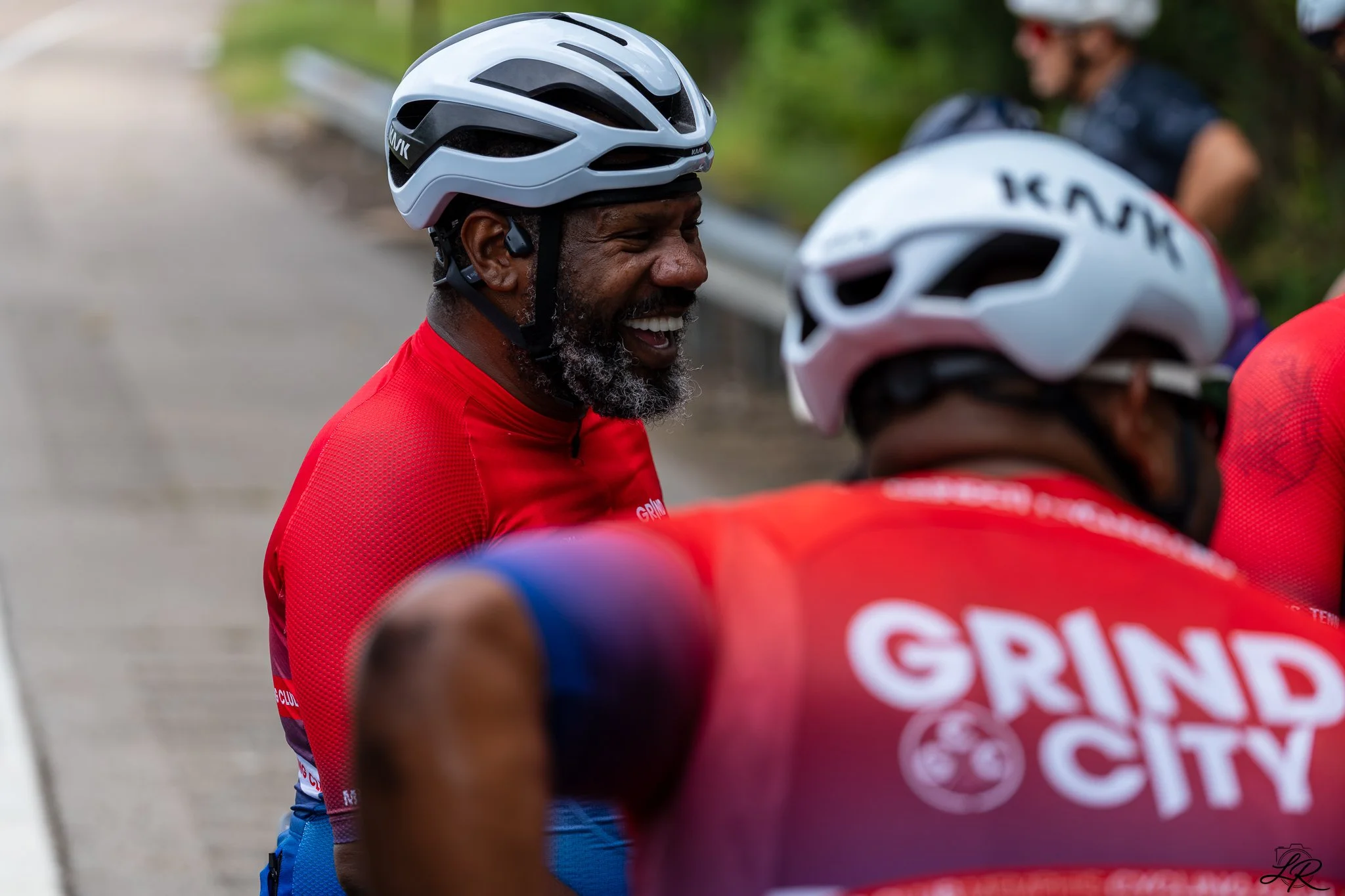 A group of cyclists wearing helmets and red jerseys, smiling and engaged in conversation during a road cycling event.