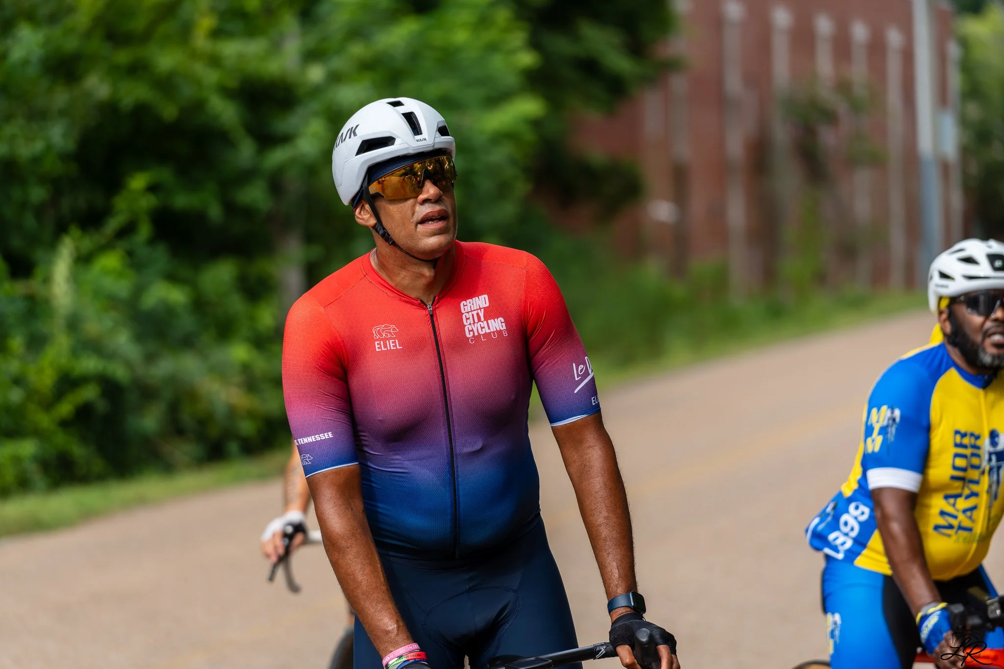 A man wearing a white helmet, sunglasses, and a red cycling jersey with purple and blue accents. He is riding a bicycle outdoors, with green trees and buildings in the background.