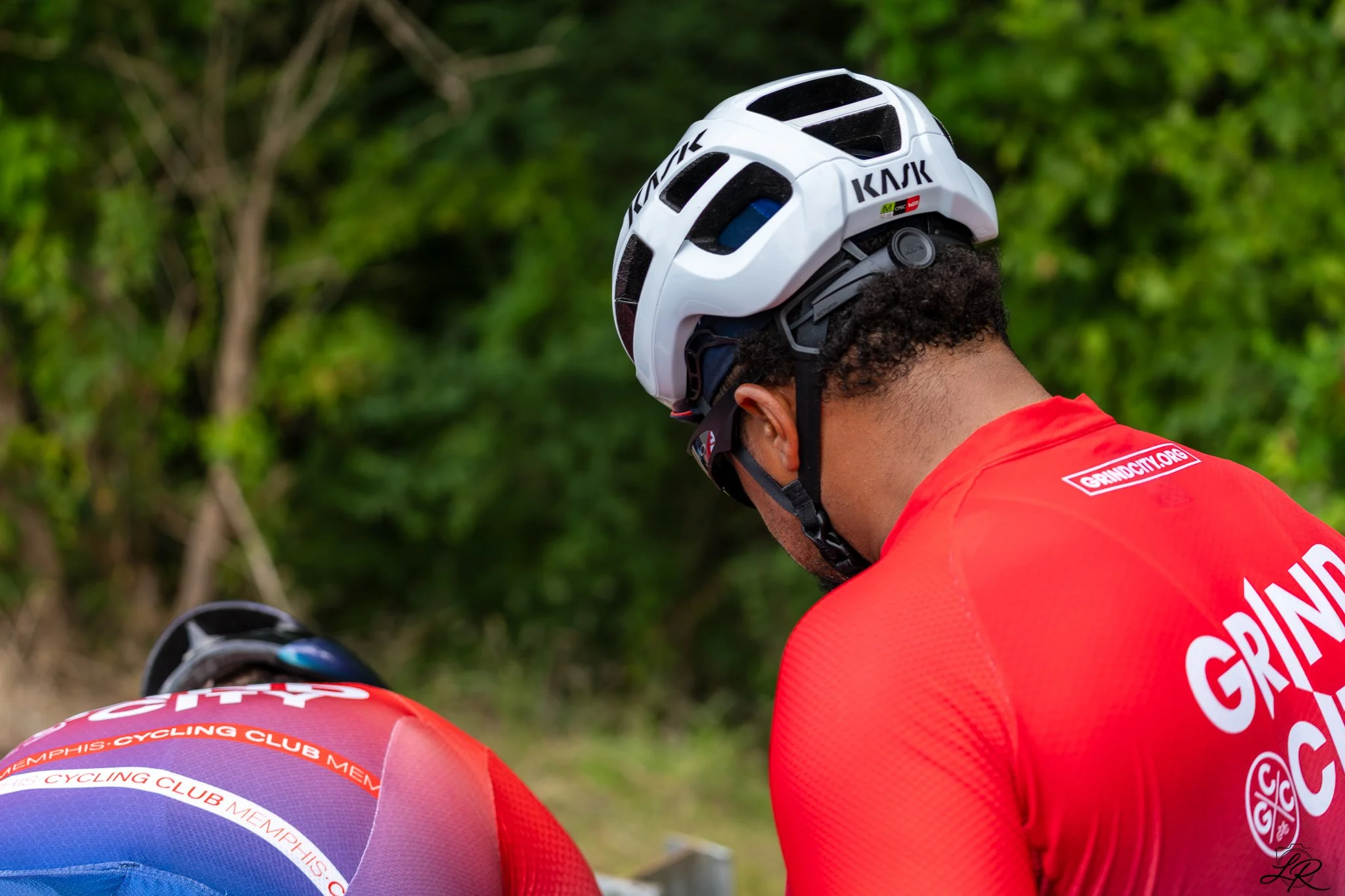 A cyclist wearing a white helmet and red jersey with Memphis cycling club branding, standing outdoors with green trees in background.