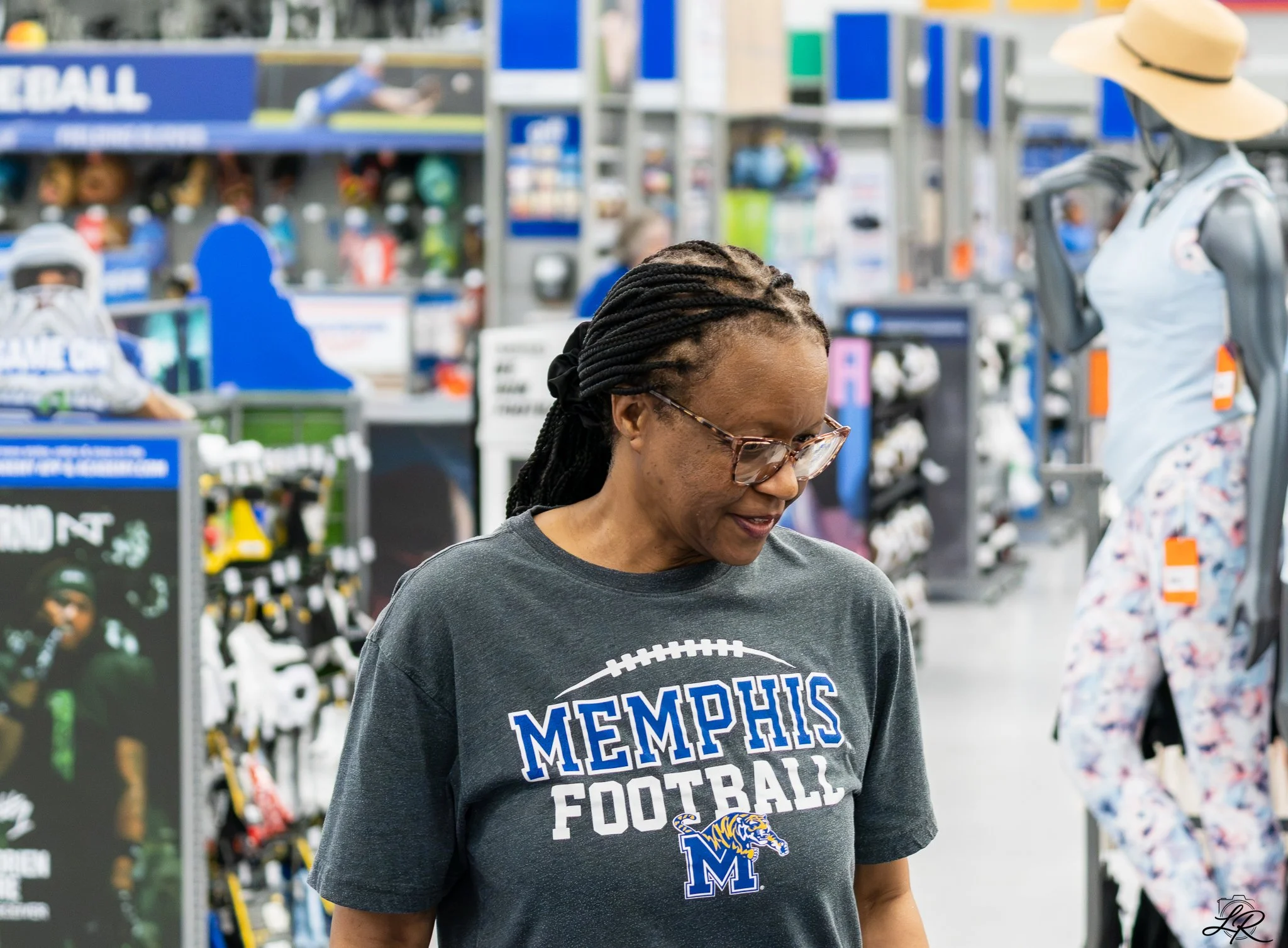 A woman with braids wearing glasses, smiling, and wearing a dark gray Memphis football T-shirt in a sports store aisle.