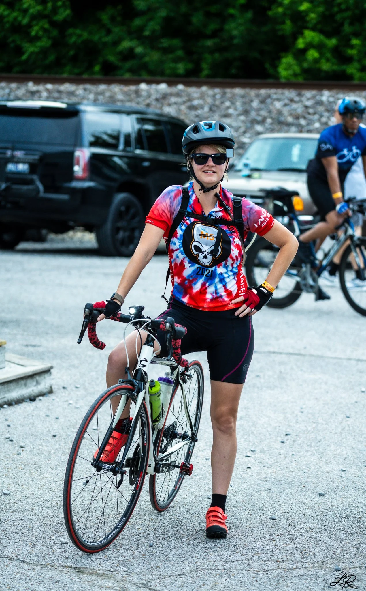 A woman in cycling gear standing next to a bicycle with a black and white frame, wearing a red, blue, and white tie-dye shirt with a skull logo, black shorts, red shoes, black helmet, and sunglasses, in a parking lot with cars and greenery in the bac