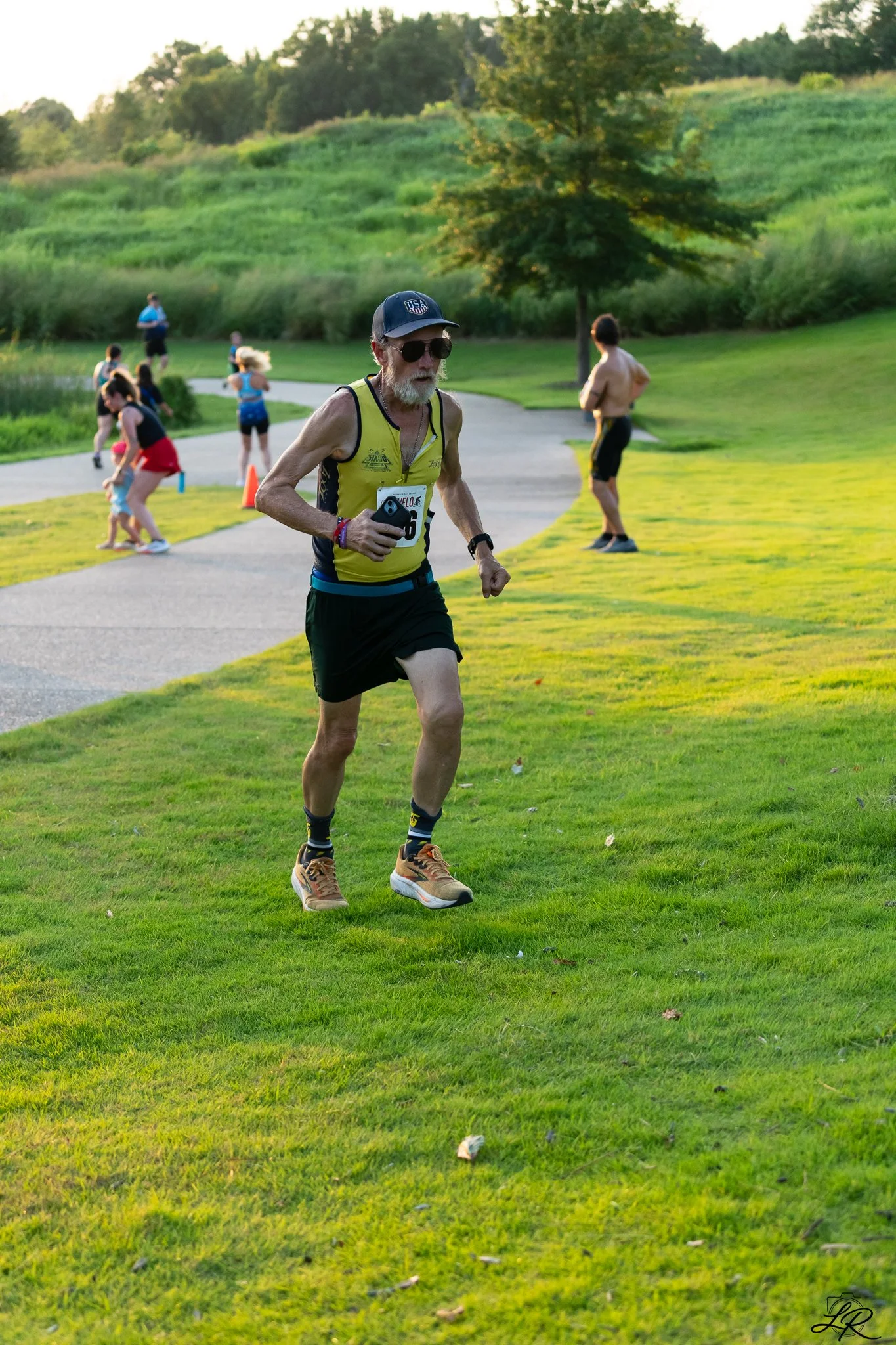 An elderly man running outdoors on a grass field during late afternoon or early evening, with people and trees in the background.