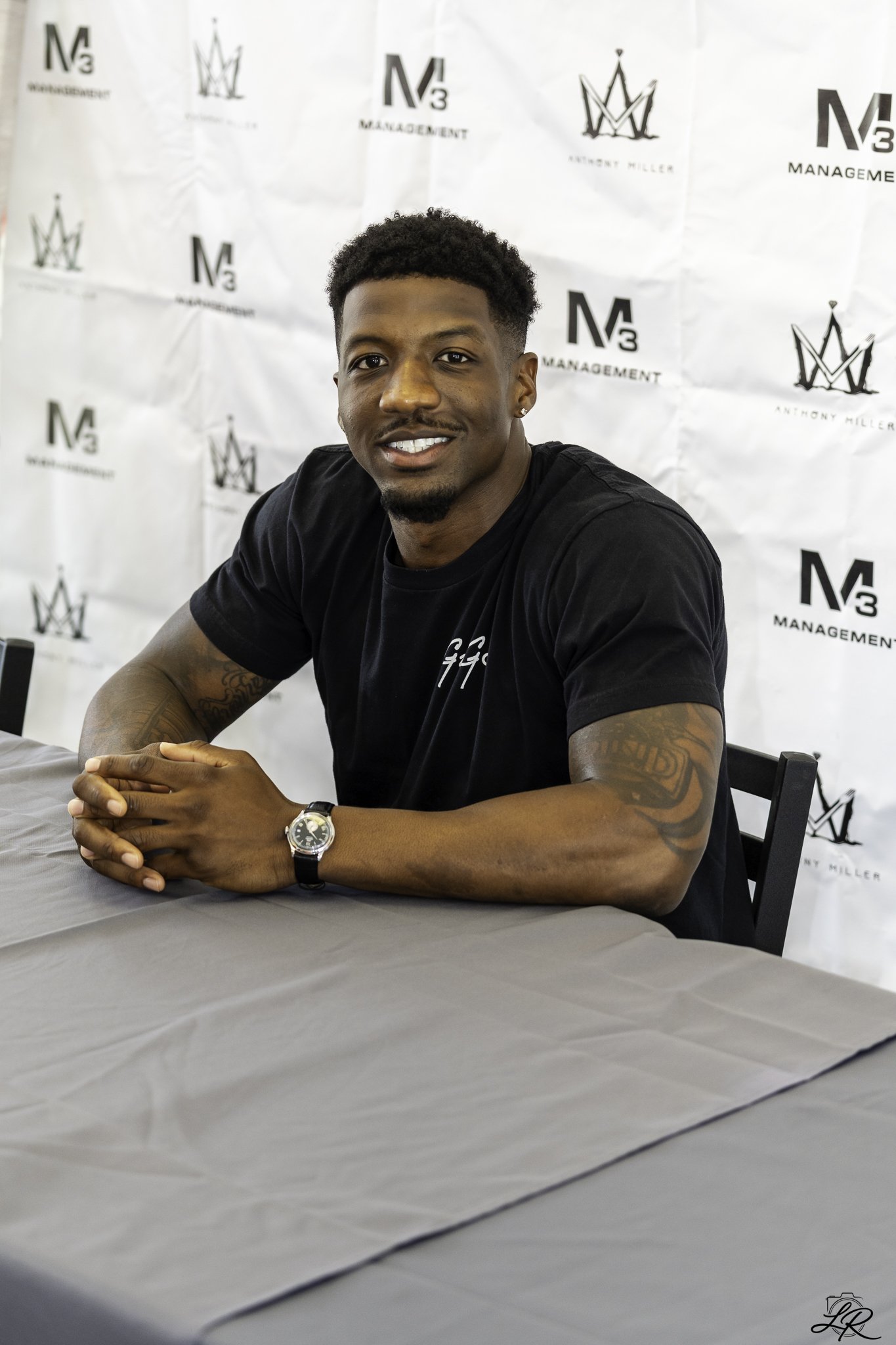 Portrait of a smiling young man with dark curly hair, wearing a black t-shirt, sitting at a table with arms folded, in front of a white backdrop with logos and text related to management.