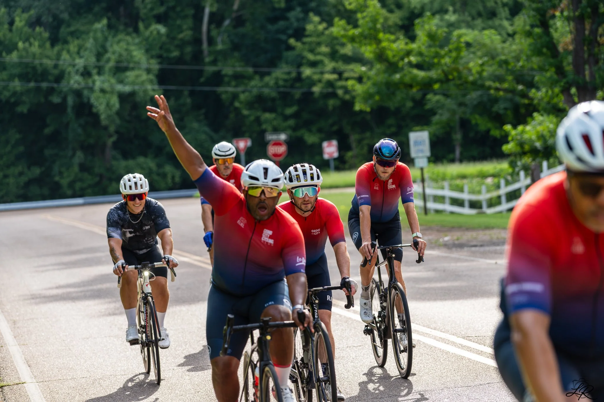 A group of cyclists riding on a road surrounded by trees, with one cyclist raising his hand.