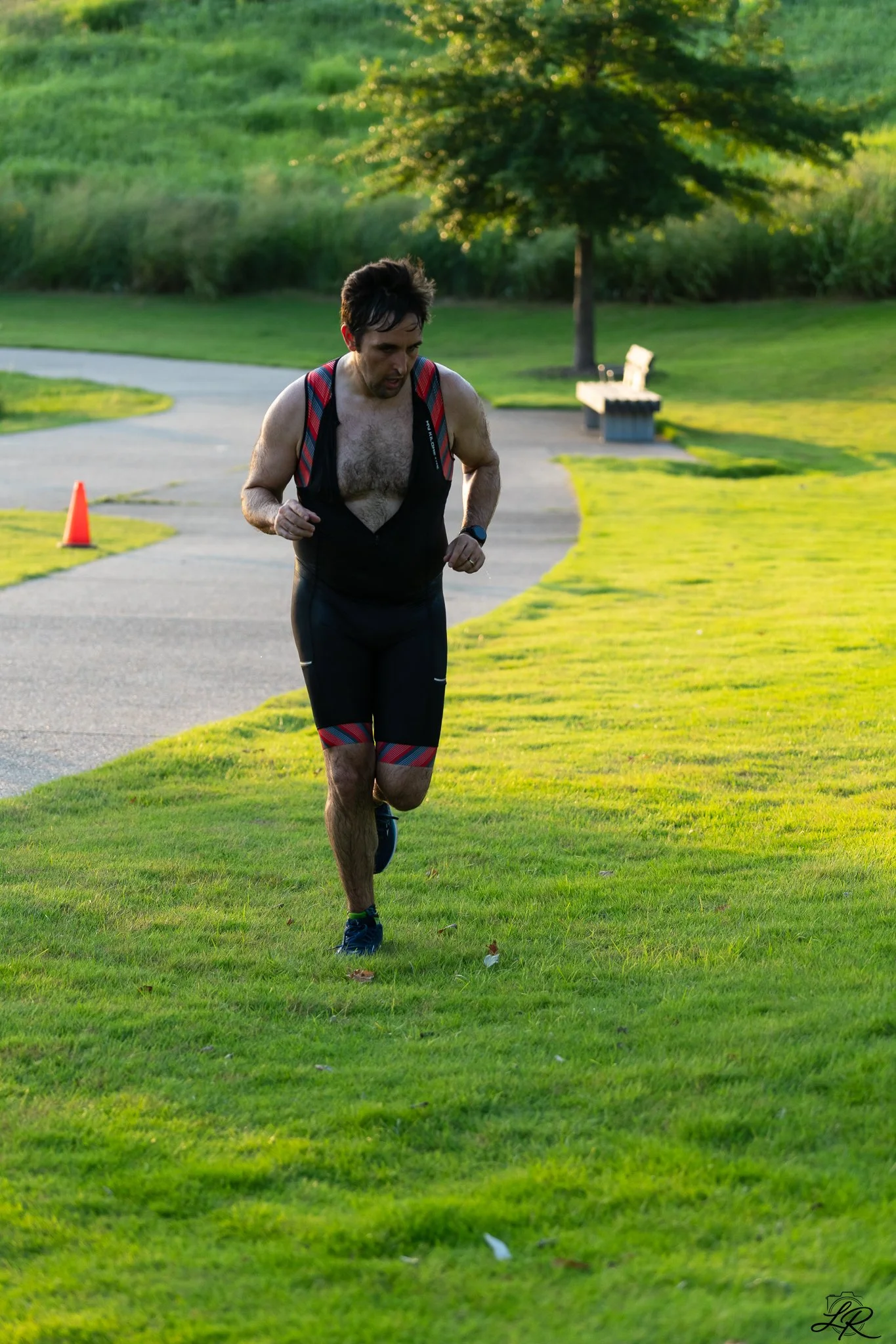 Man running on a grassy park with a backpack and a watch, with trees and a bench in the background.