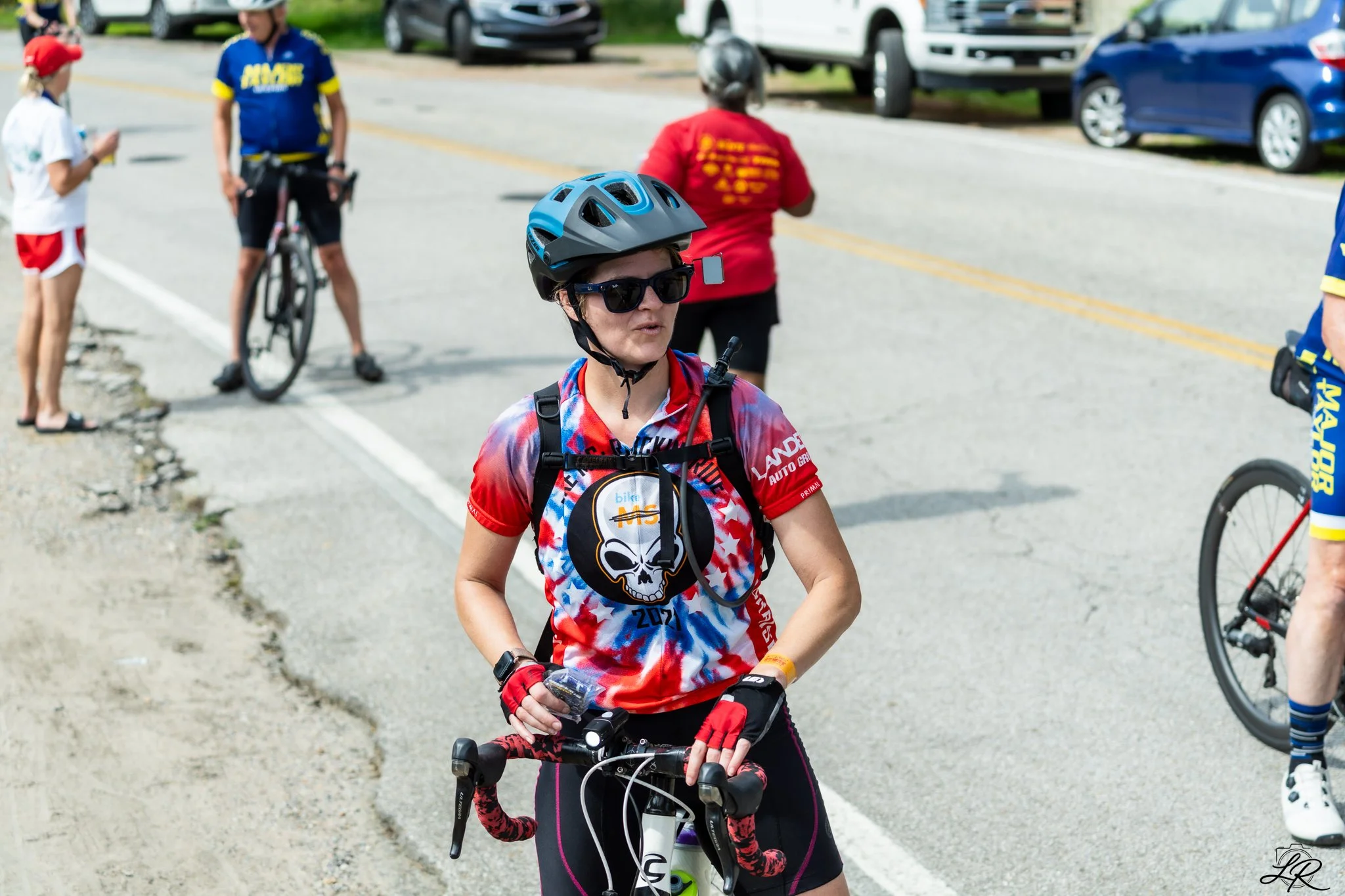A woman with blonde hair, wearing black sunglasses, a black and red helmet, and a colorful cycling jersey with a skull design, stands with her bike. She is surrounded by other cyclists and spectators on a street, with parked cars and a grassy area in